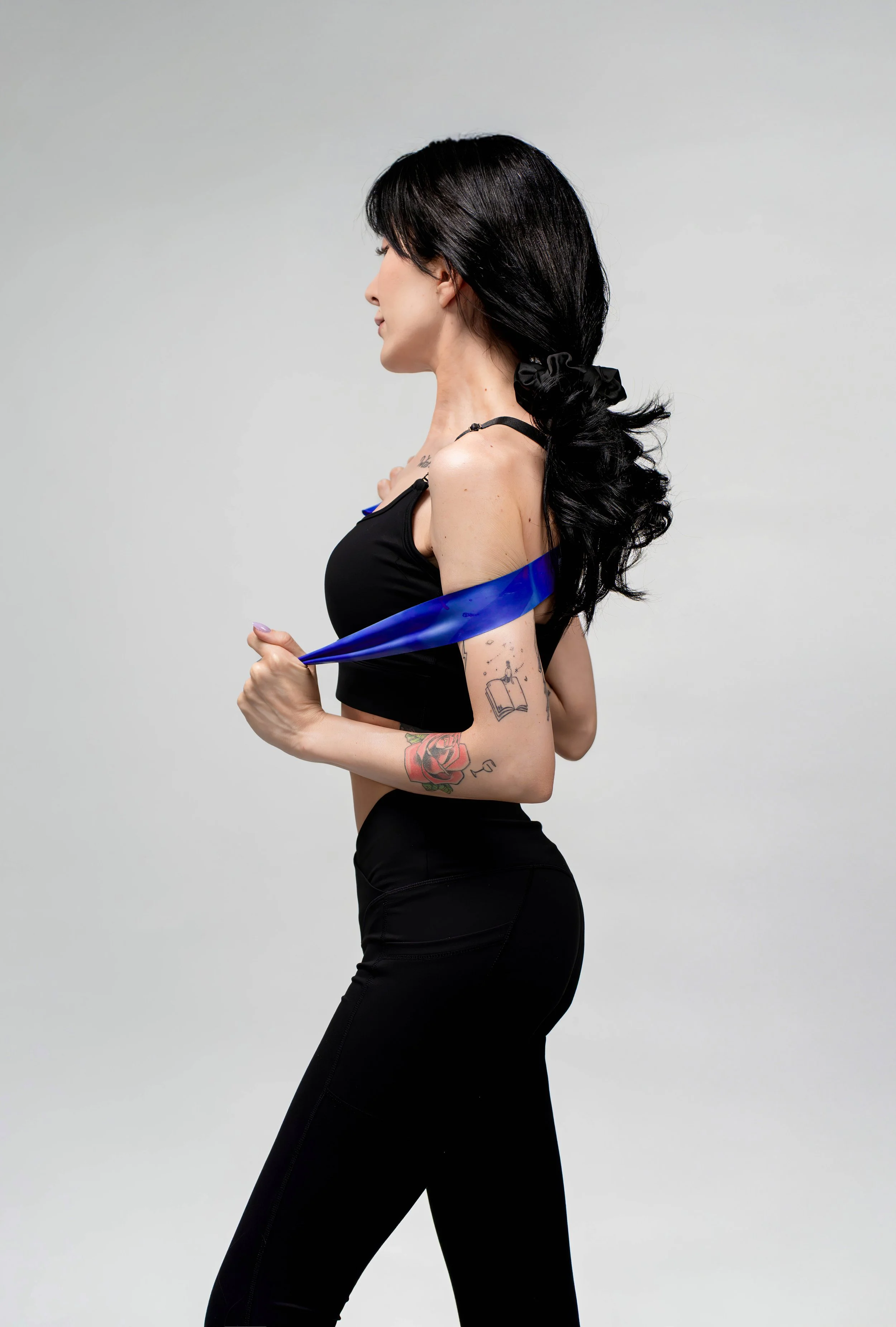 A woman with long black hair, wearing black athletic clothing, pulls a blue resistance band across her shoulders, standing against a plain light gray background.