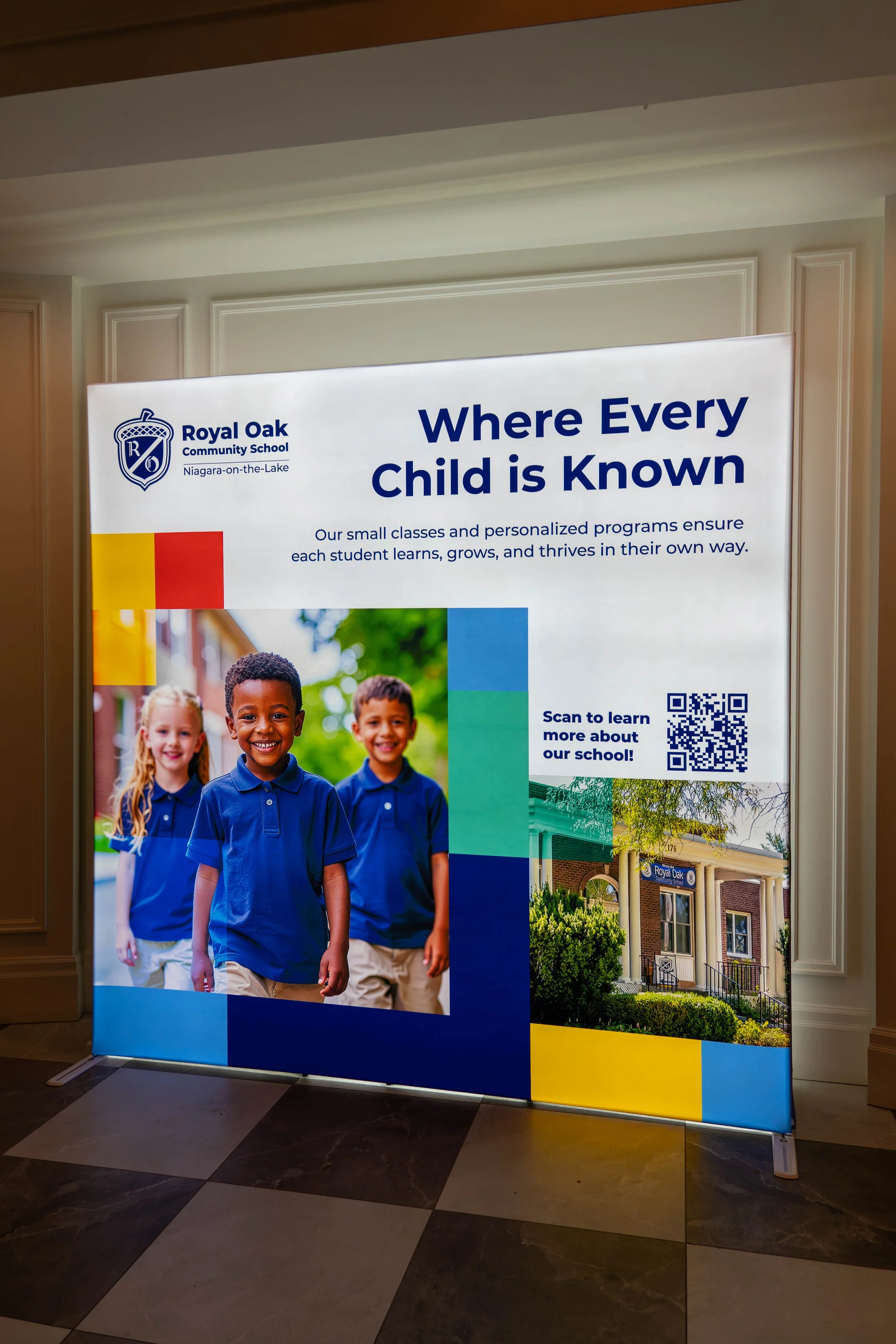 A promotional display for Royal Oak Community School Niagara-on-the-Lake, showing a smiling group of children in blue uniforms, a QR code to learn more, and an image of the school building.