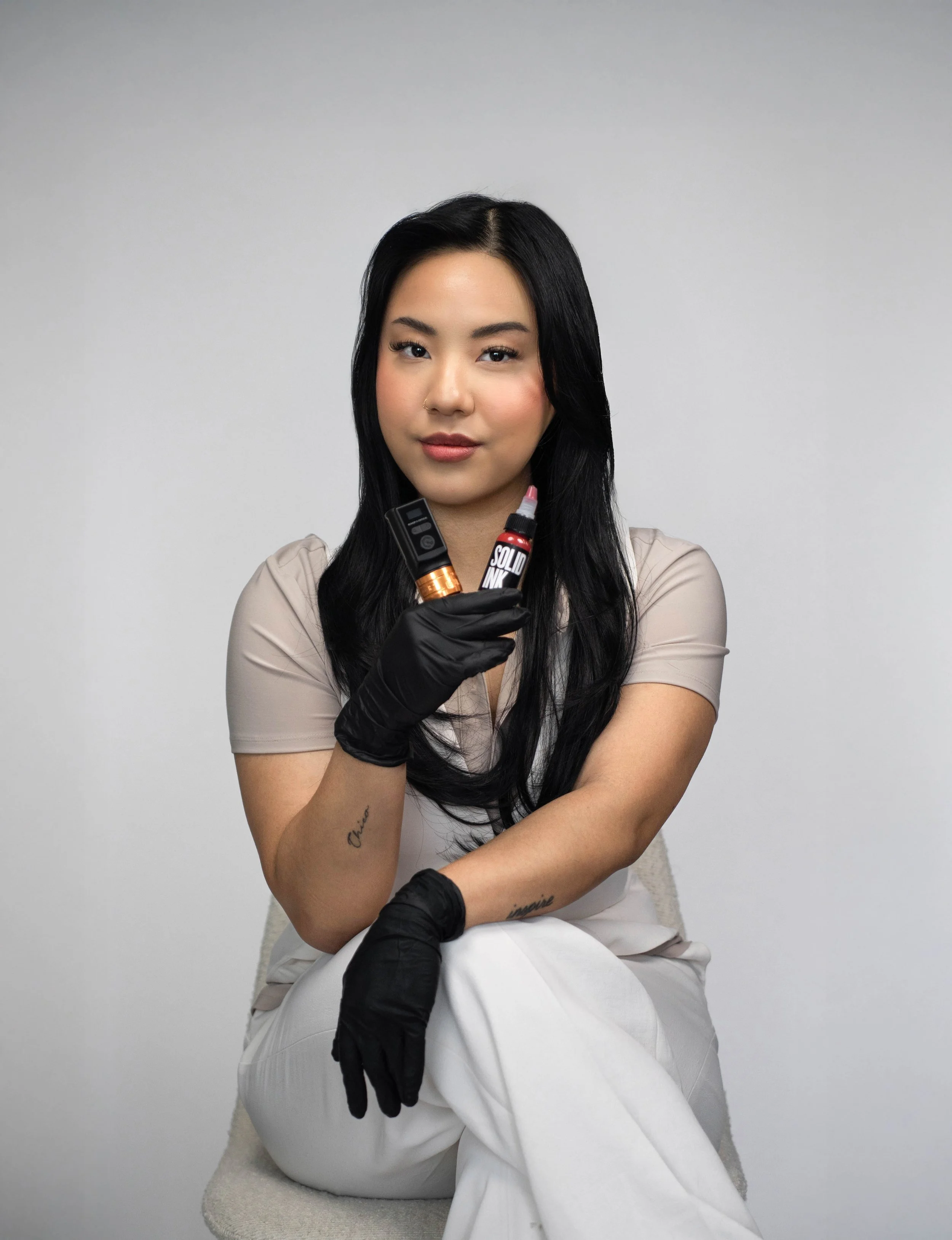 A woman with long dark hair and light skin sits on a white chair against a plain background. She wears a beige top, white pants, and black gloves. She holds tattoo equipment including a tattoo machine and ink bottles, and looks confidently at the cam