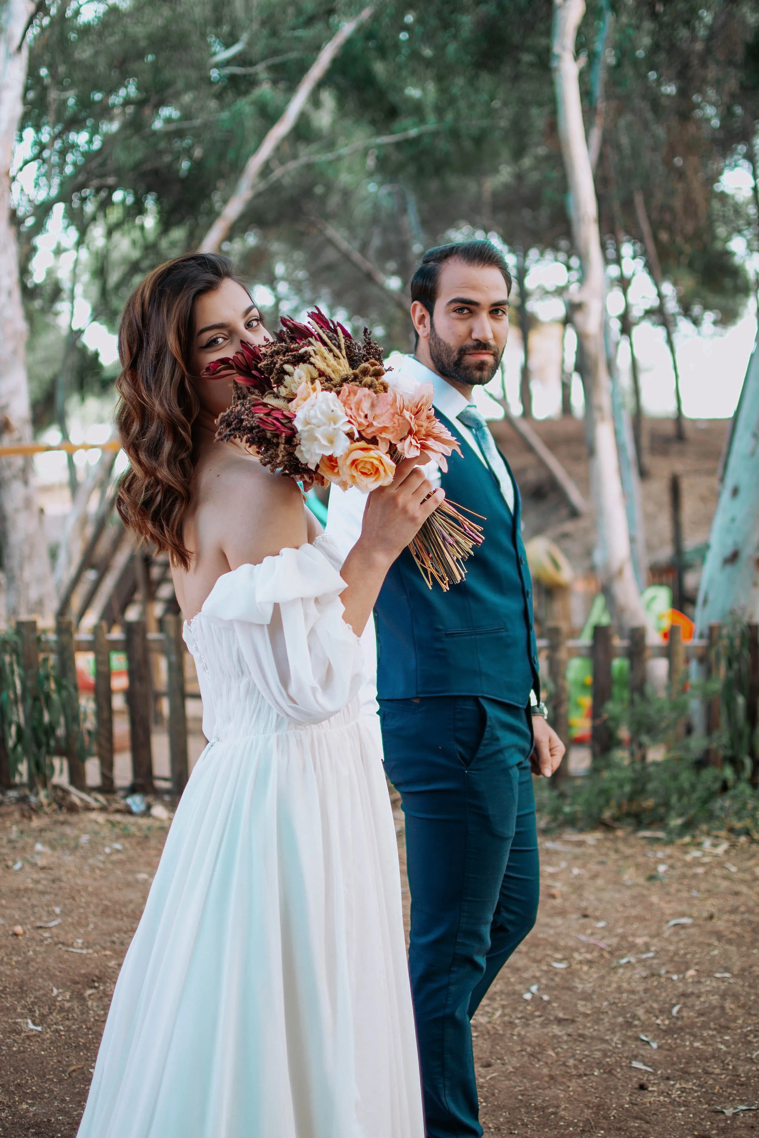 A woman in a white off-the-shoulder dress holding a bouquet of flowers, standing outdoors with a man in a navy blue suit and tie, trees in the background.
