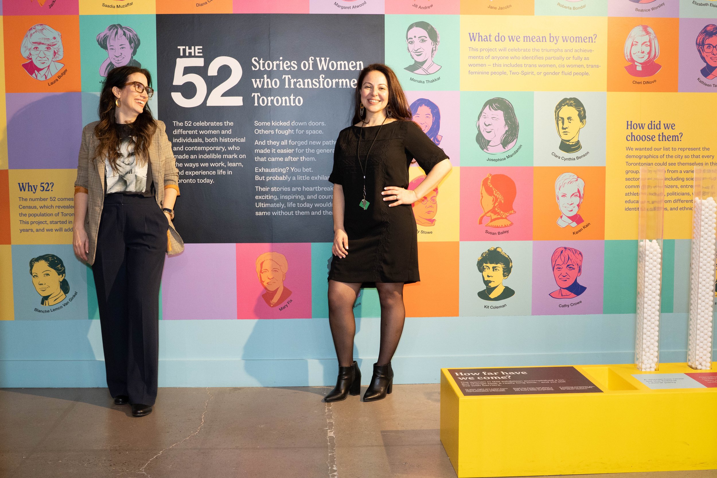 Two women standing in front of a colorful mural titled 'The 52 Stories of Women who Transformed Toronto,' featuring illustrations and names of various women, discussing the project celebrating women and gender identities.