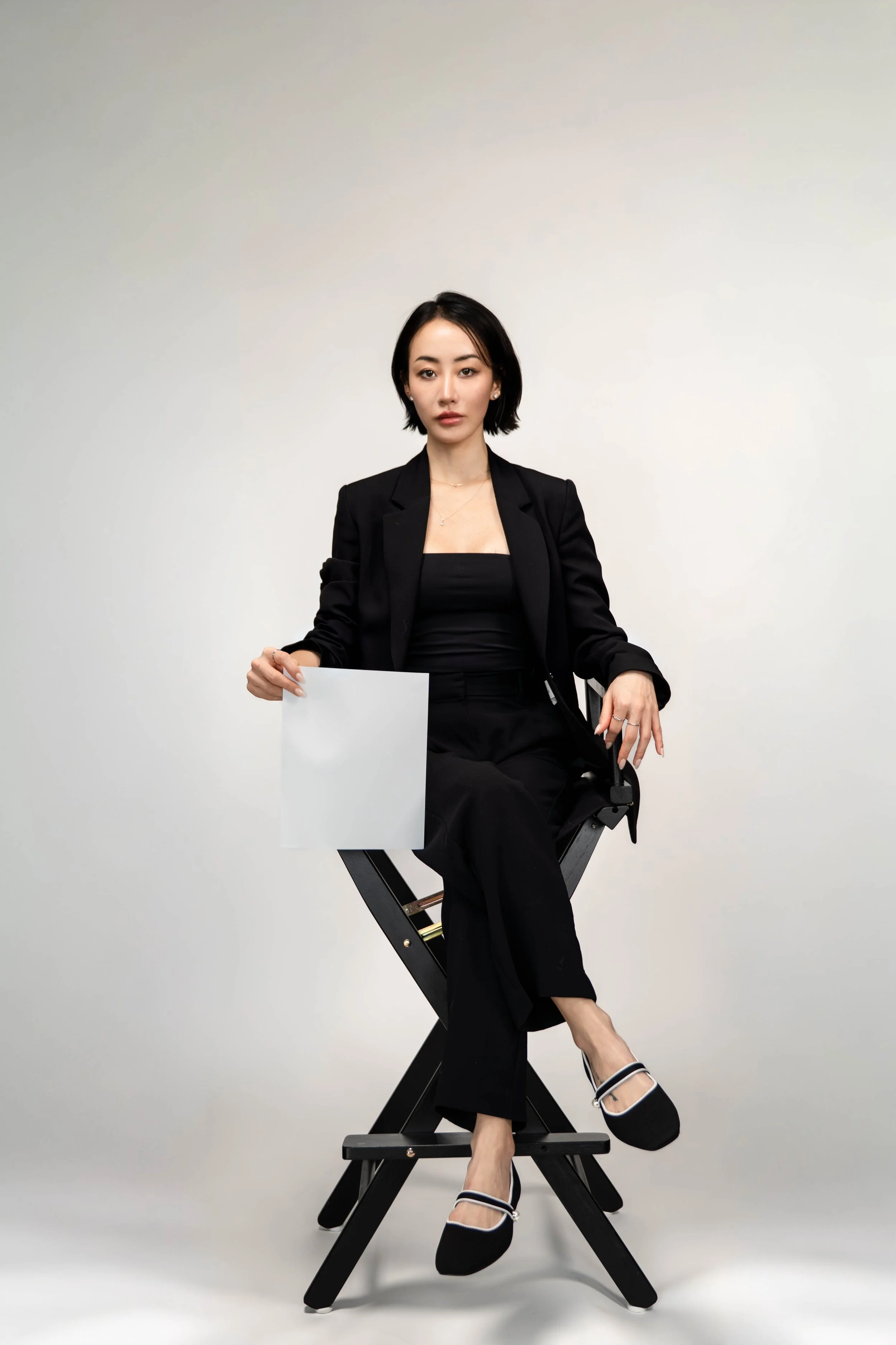 A woman in a black blazer, black top, and black pants sitting on a director's chair against a plain white background, holding a blank sheet of paper.