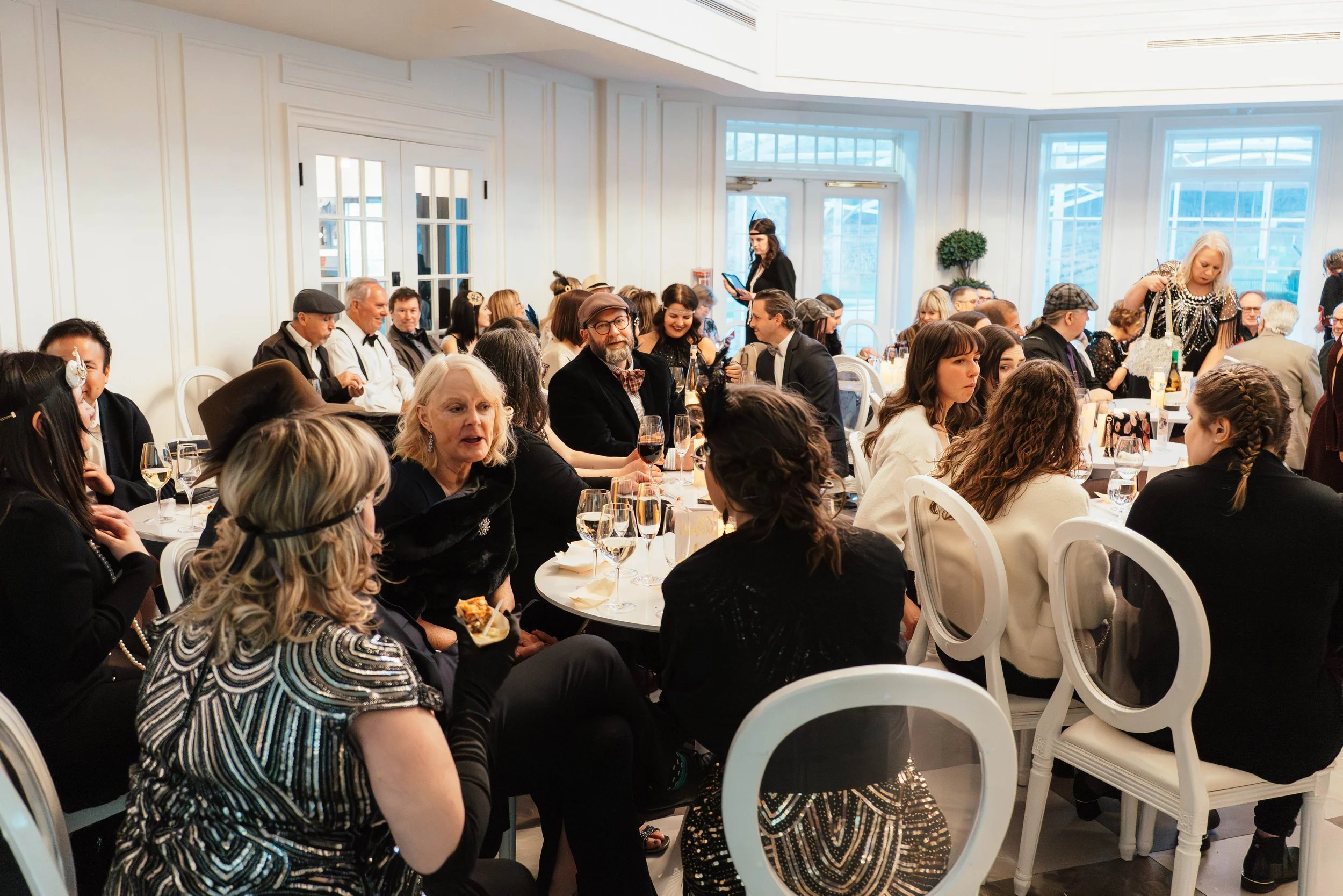 People seated at round tables in a well-lit room with large windows, attending a social event or party, some holding glasses of wine.