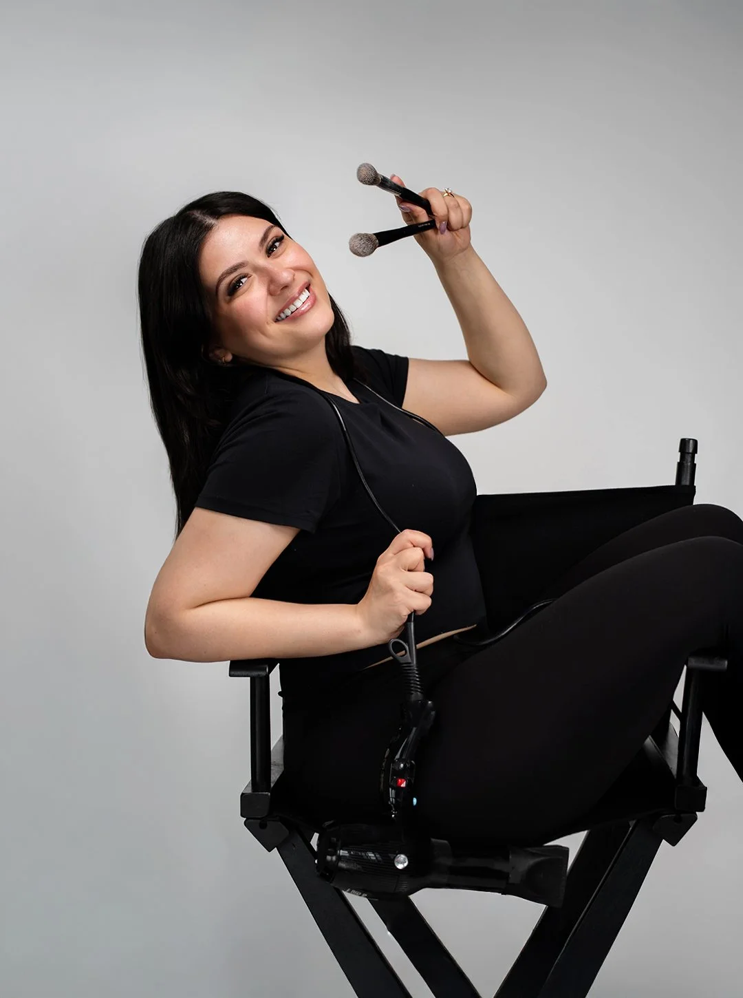 Woman with dark hair, smiling, sitting in an electric wheelchair, holding makeup brushes, wearing black clothing, white background.