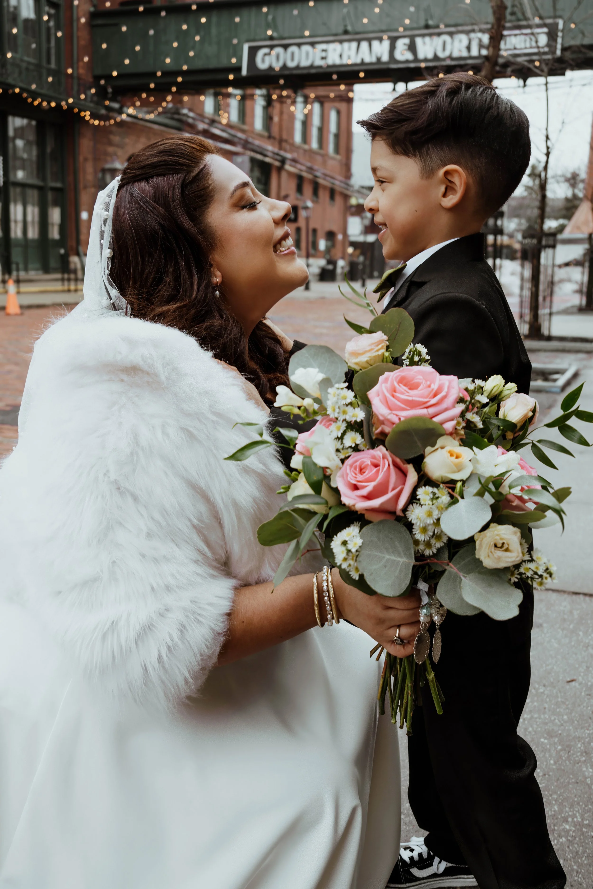 A bride and young boy sharing a joyful moment, with the bride holding a bouquet of pink roses and white flowers, in an outdoor urban setting