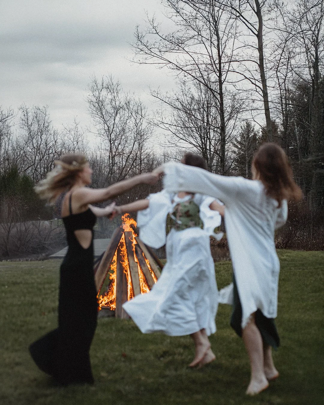 Three women dancing in front of a bonfire during dusk in a backyard with leafless trees.