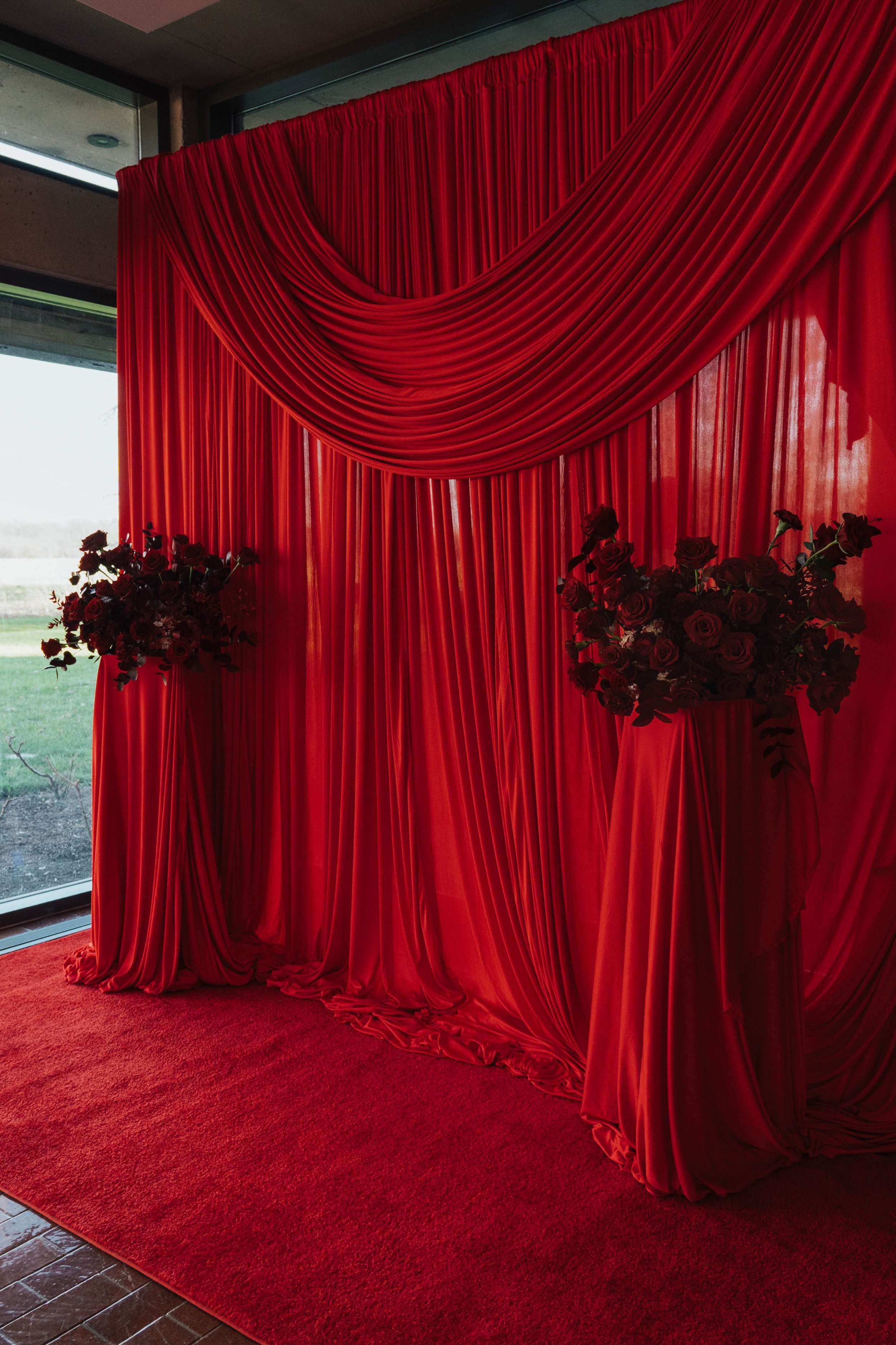 Red draped fabric backdrop with floral arrangements on pedestals at a decorated event space