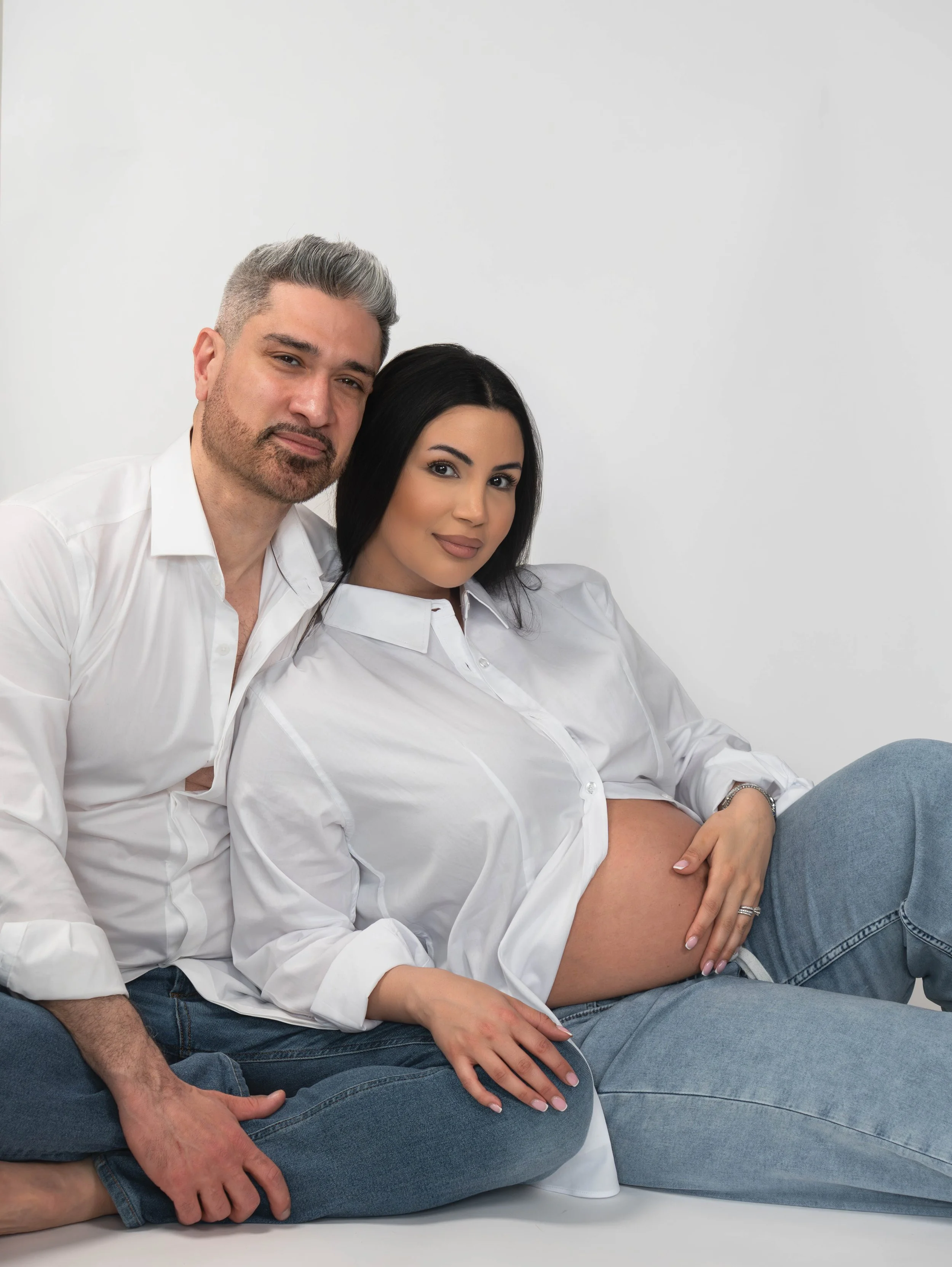 A man and woman sitting on a white surface against a plain white background. The man has gray hair, a beard, and is wearing a white shirt with rolled-up sleeves and blue jeans. The woman has long dark hair, wearing a white blouse and blue jeans, with