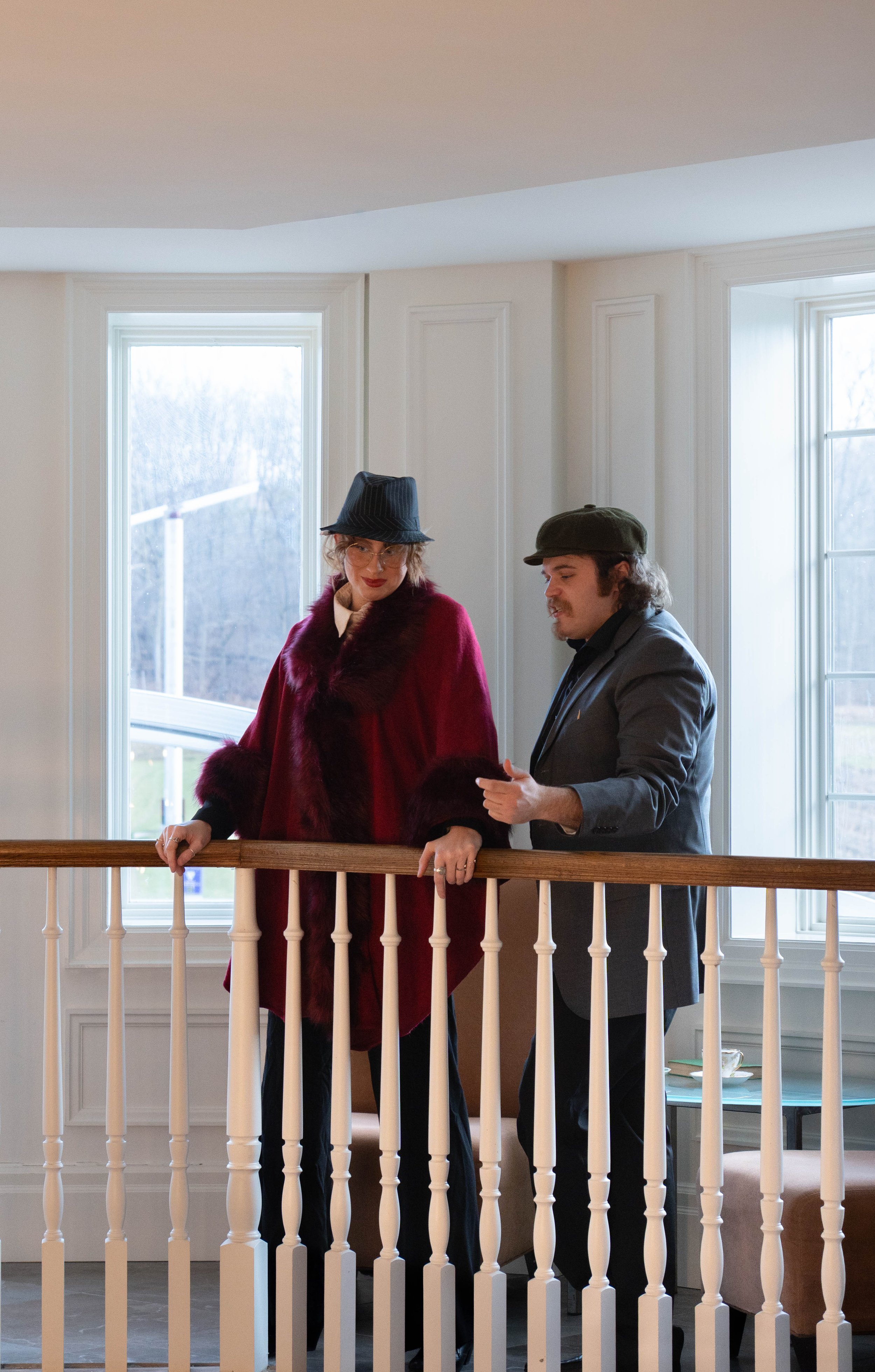 A man and woman stand on a staircase landing inside a house, looking out the window. The woman is wearing a dark blue hat, red coat with fur trim, and glasses. The man is wearing a cap and gray jacket, and appears to be holding a phone or small devic