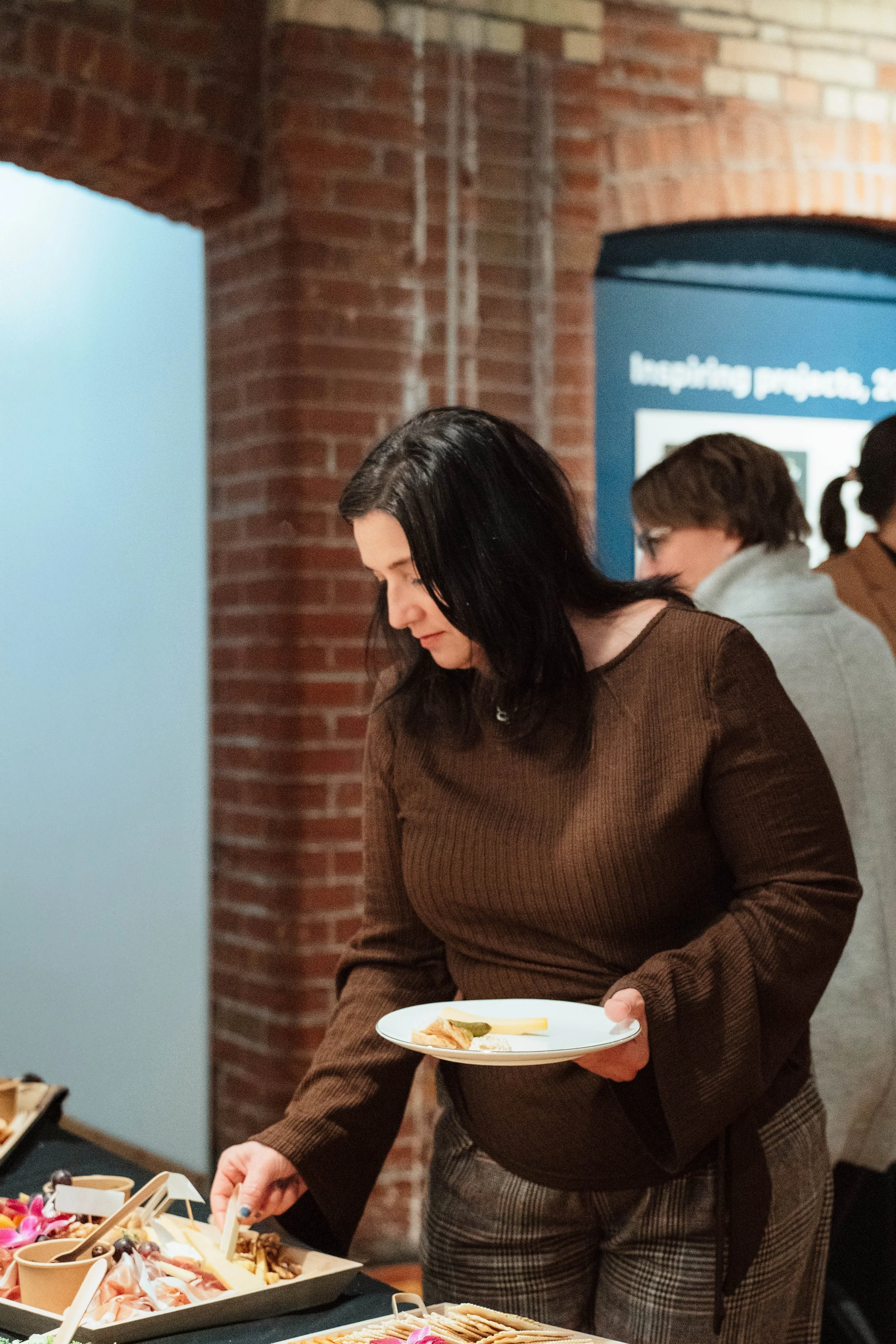A woman serving herself at a buffet table with various food options, standing in front of a brick wall.