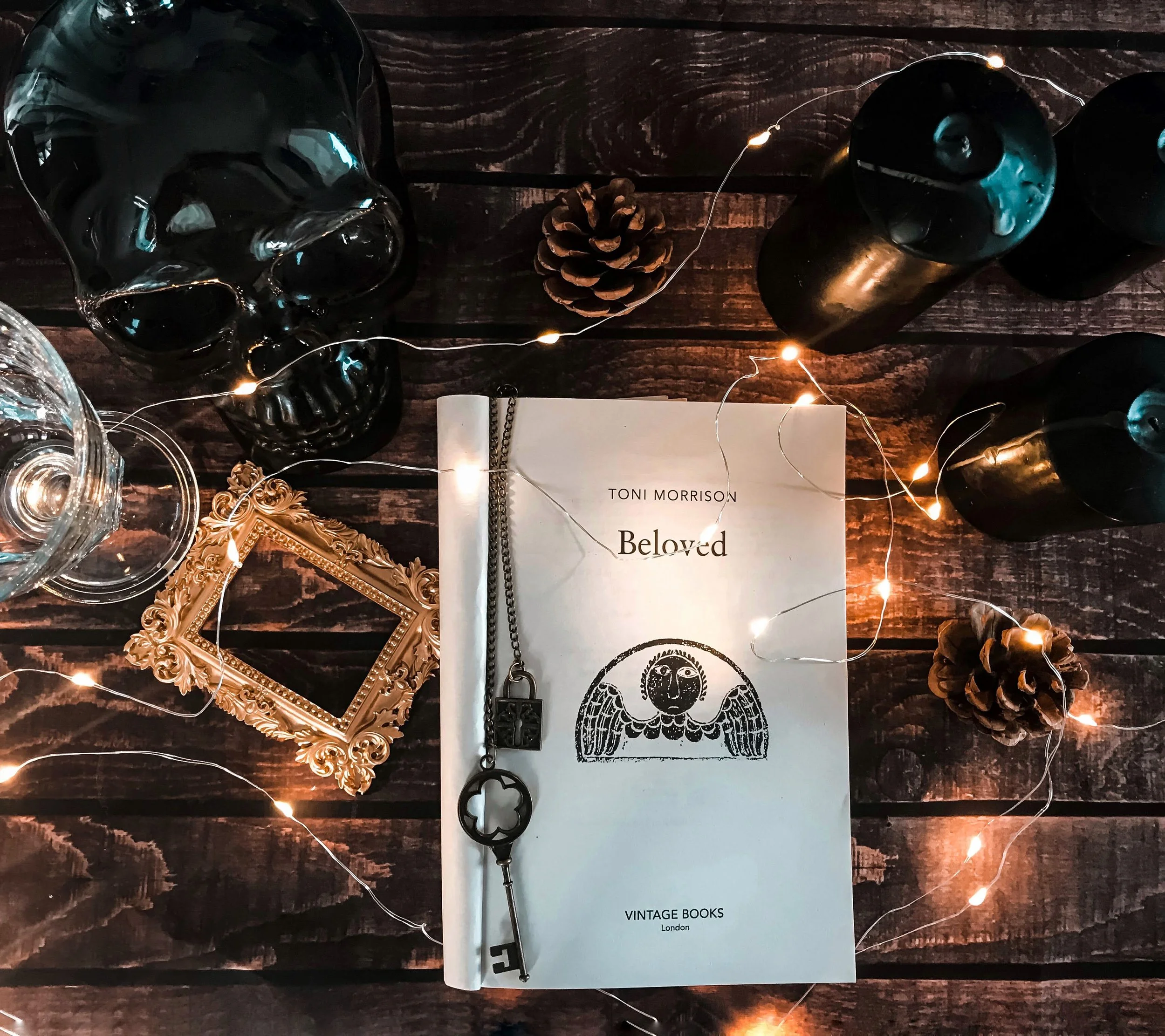 A flat lay of a dark wooden table with various items: a skull-shaped black glass container, a gold ornate picture frame, a glass decanter, pine cones, a book titled 'Beloved' by Toni Morrison, a string of fairy lights, black candles, and vintage-styl