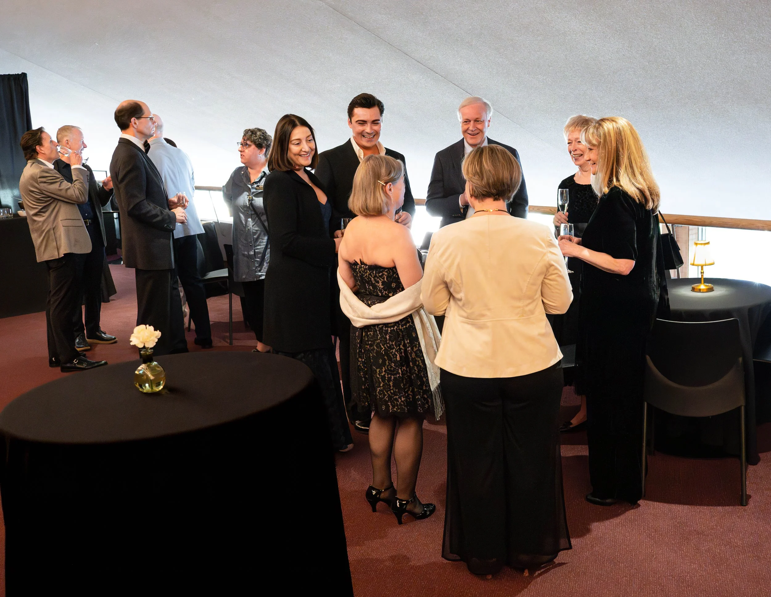 Group of people at a social gathering, engaging in conversation, with some holding drinks, in a room with a sloped ceiling and round tables