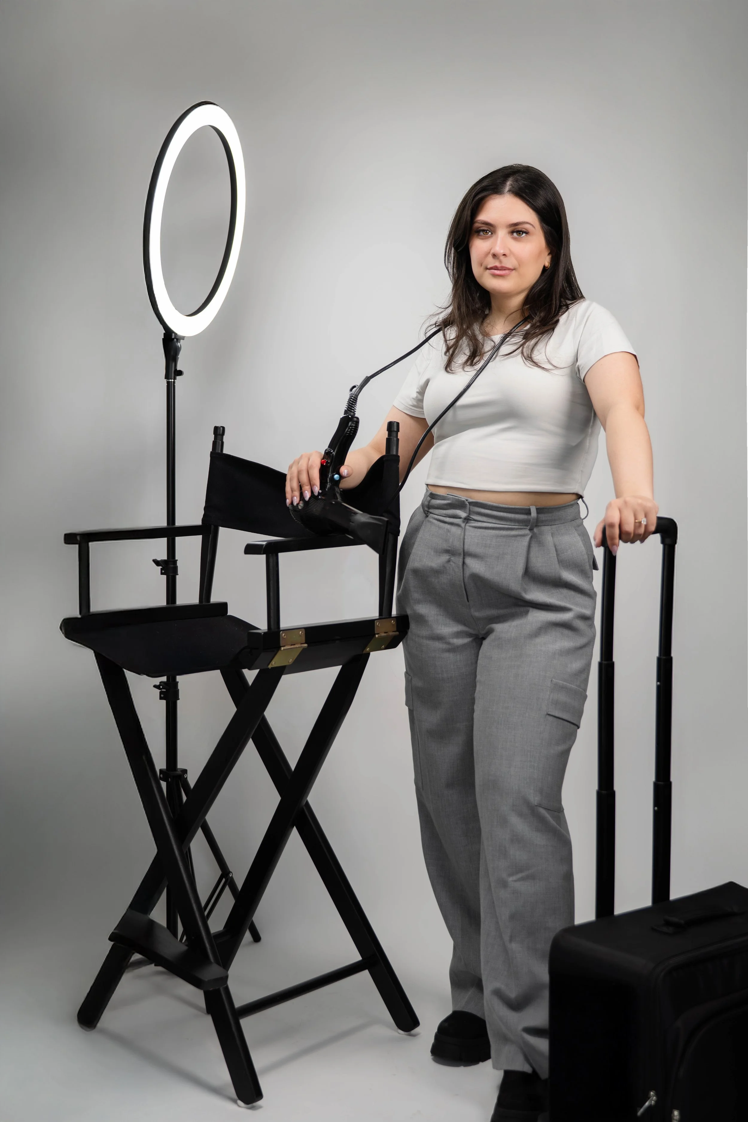 Woman standing next to a director's chair and rolling suitcase, holding a remote and sitting in front of a ring light.