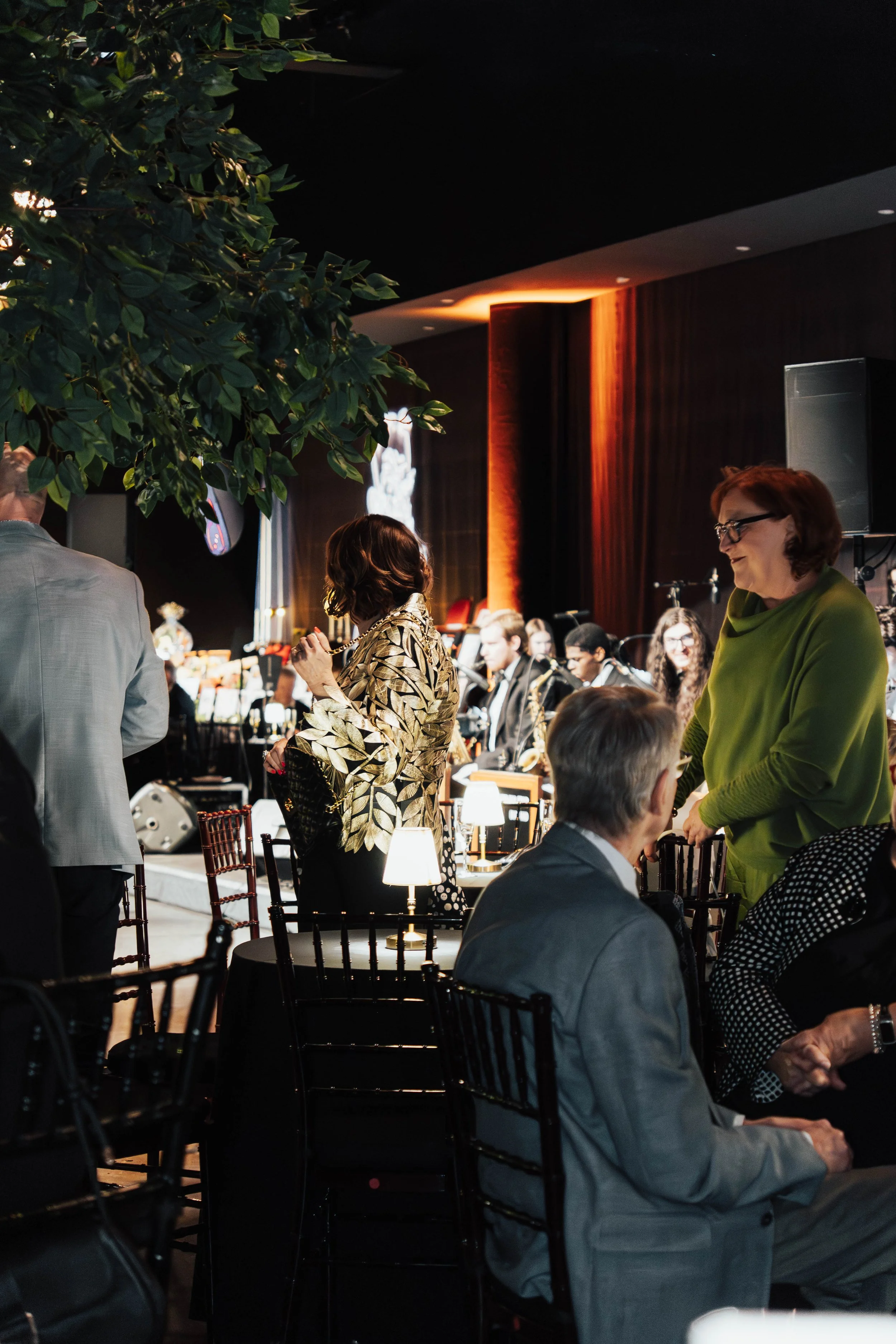 People attending a formal event with an orchestra performing on stage in the background, in a dimly lit room with elegant decor.
