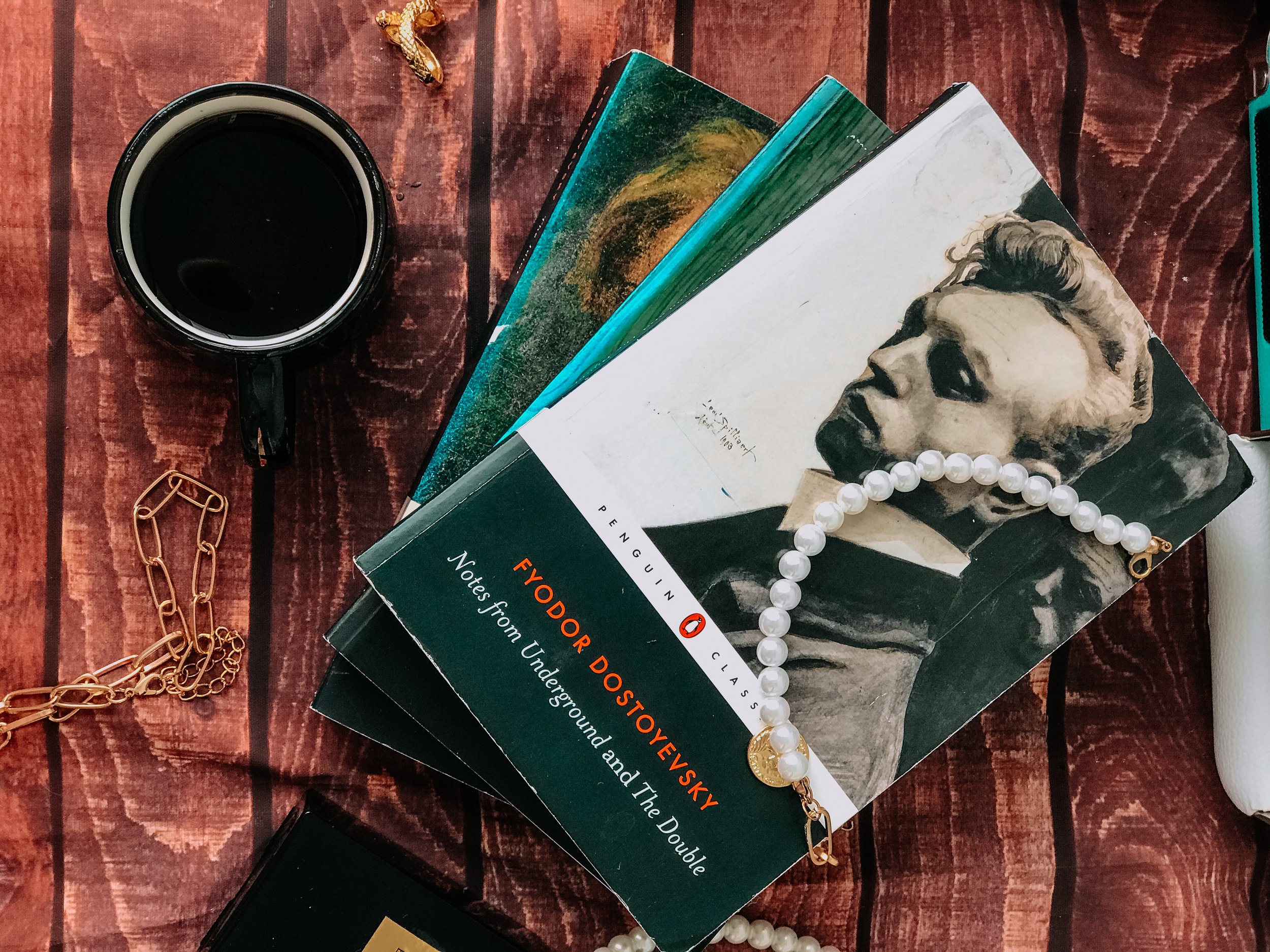 A wooden table with a black coffee mug, a gold chain necklace, a book titled "Notes from Underground and The Double" by Fyodor Dostoevsky, and a pearl necklace.