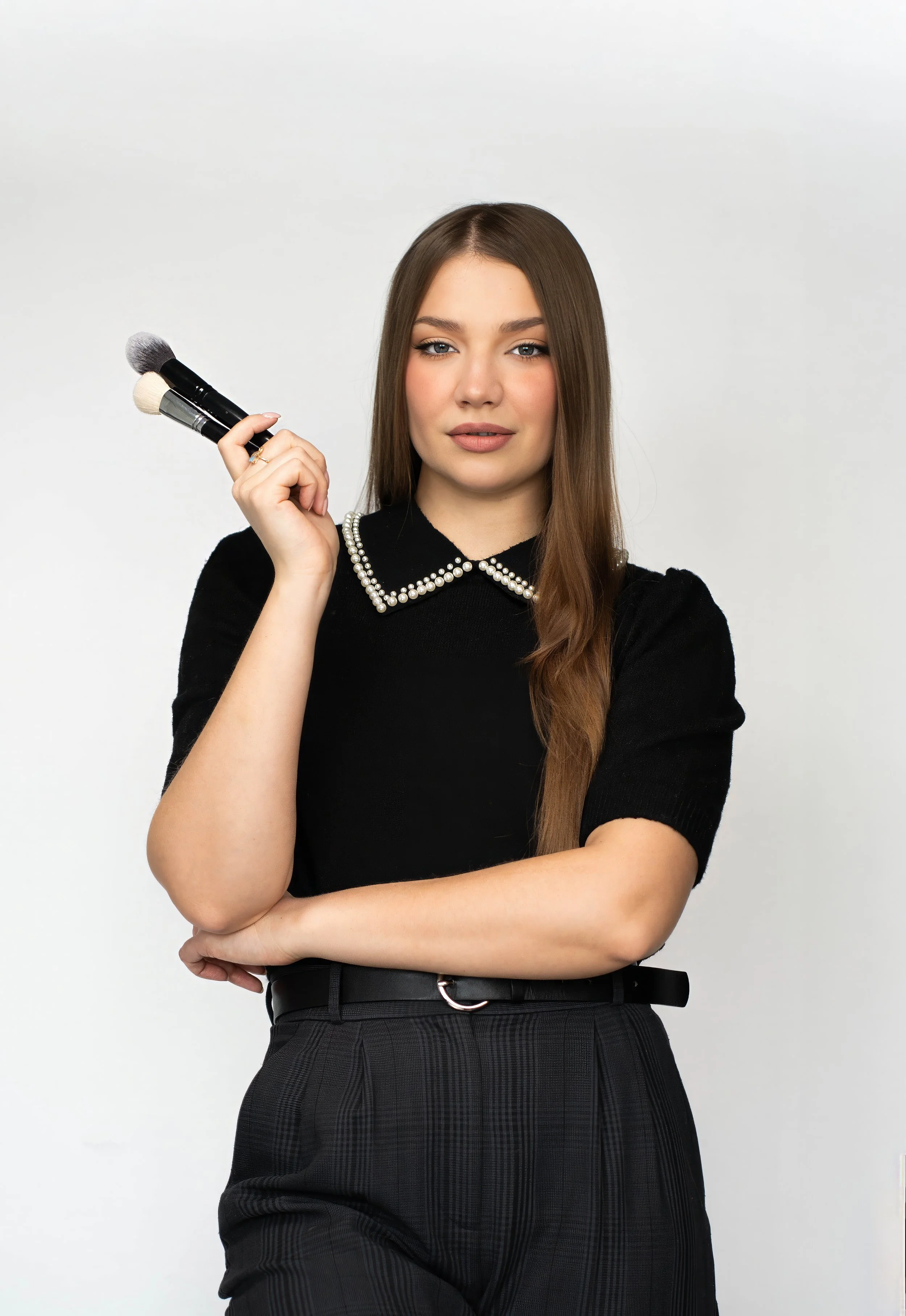 A young woman with long brown hair holds makeup brushes and looks at the camera against a white background. She wears a black top with a pearl-embellished collar and plaid pants.