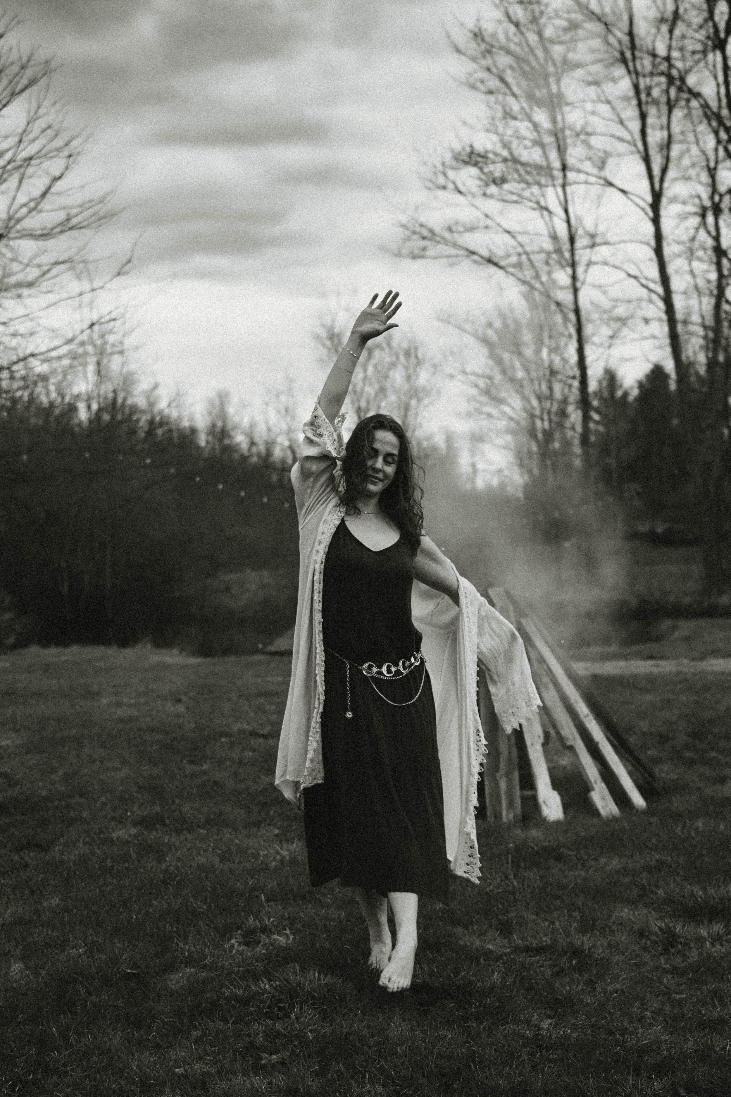A woman in a flowing dress and cardigan walking barefoot outdoors, smiling, with trees and a cloudy sky in the background.