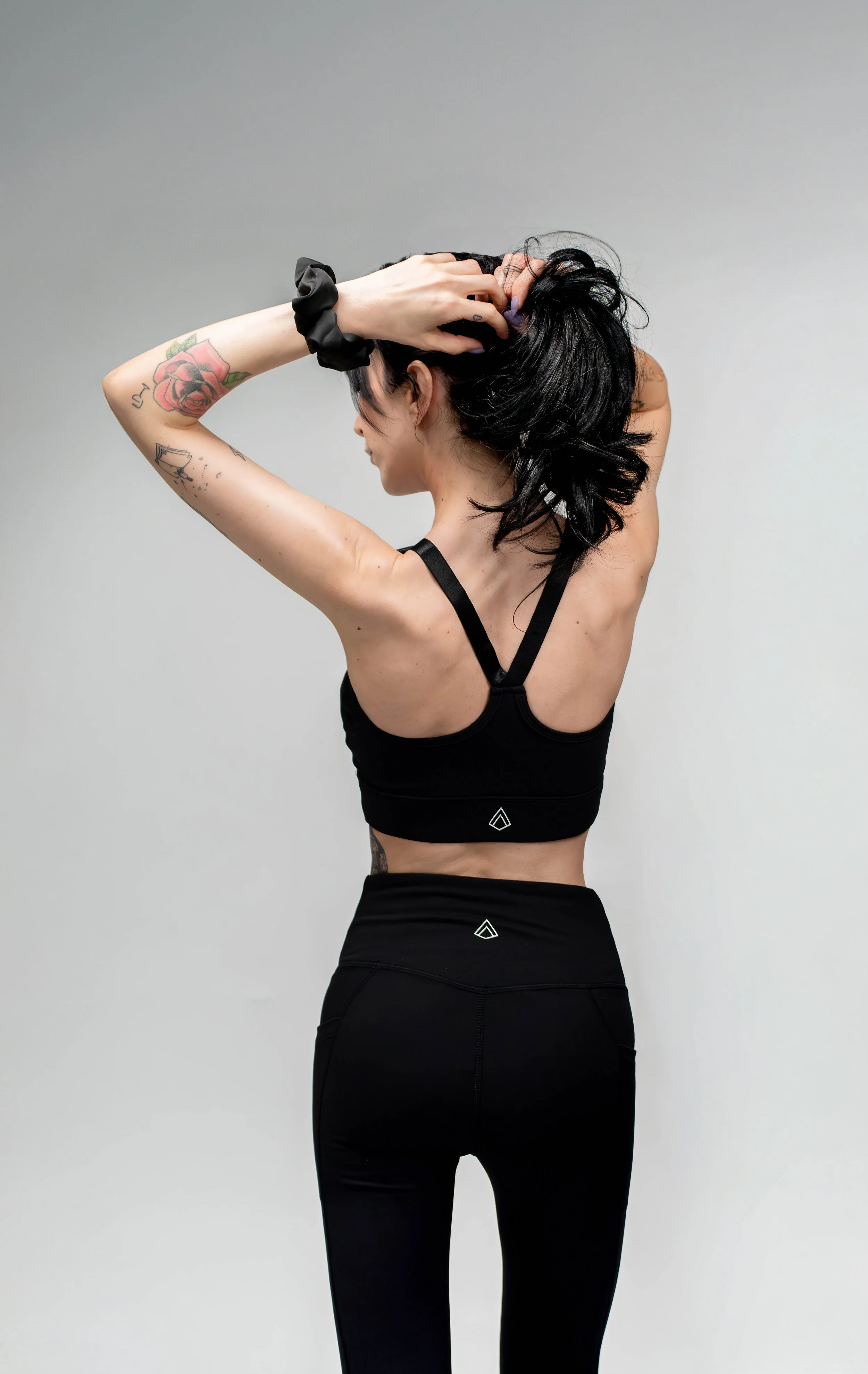 A woman with black hair wearing black sportswear, stretching with her hands in her hair, standing against a plain white background.