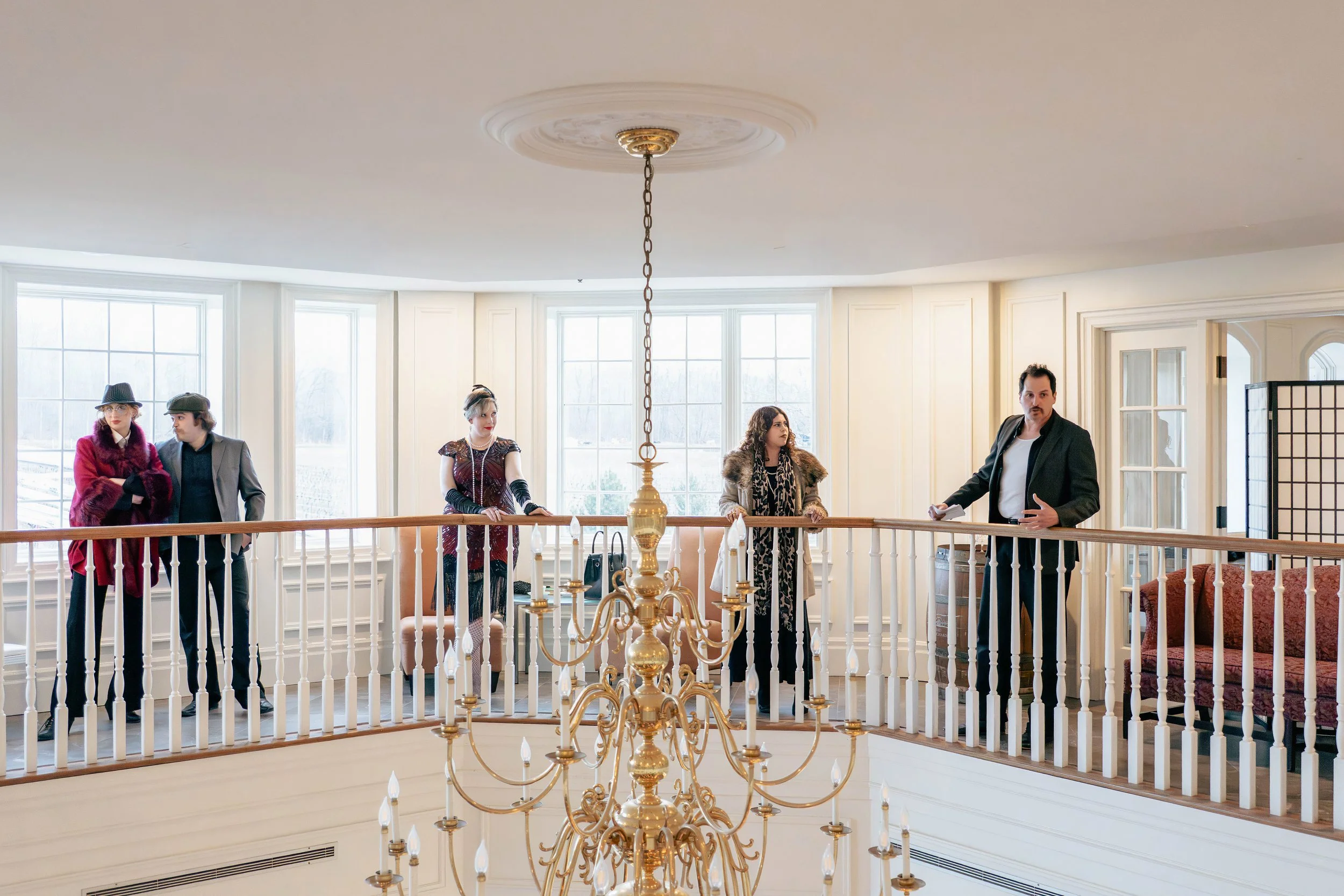 Five people stand on a balcony with large windows in the background. The group includes four women and one man, dressed in vintage or costume clothing, with a chandelier hanging from the ceiling in front of them.
