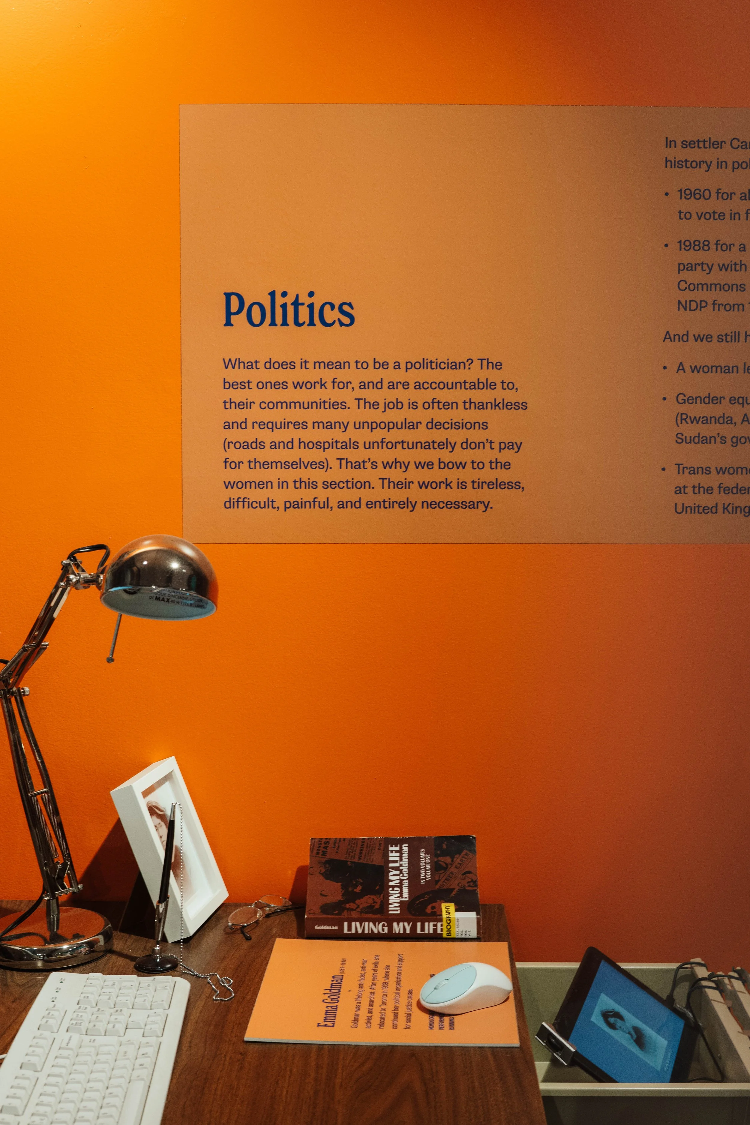 A workspace with a computer keyboard, a framed photo, glasses, a book titled "Living My Life," an orange notepad, a white computer mouse, a desk lamp, and an informational poster about politics on an orange wall.