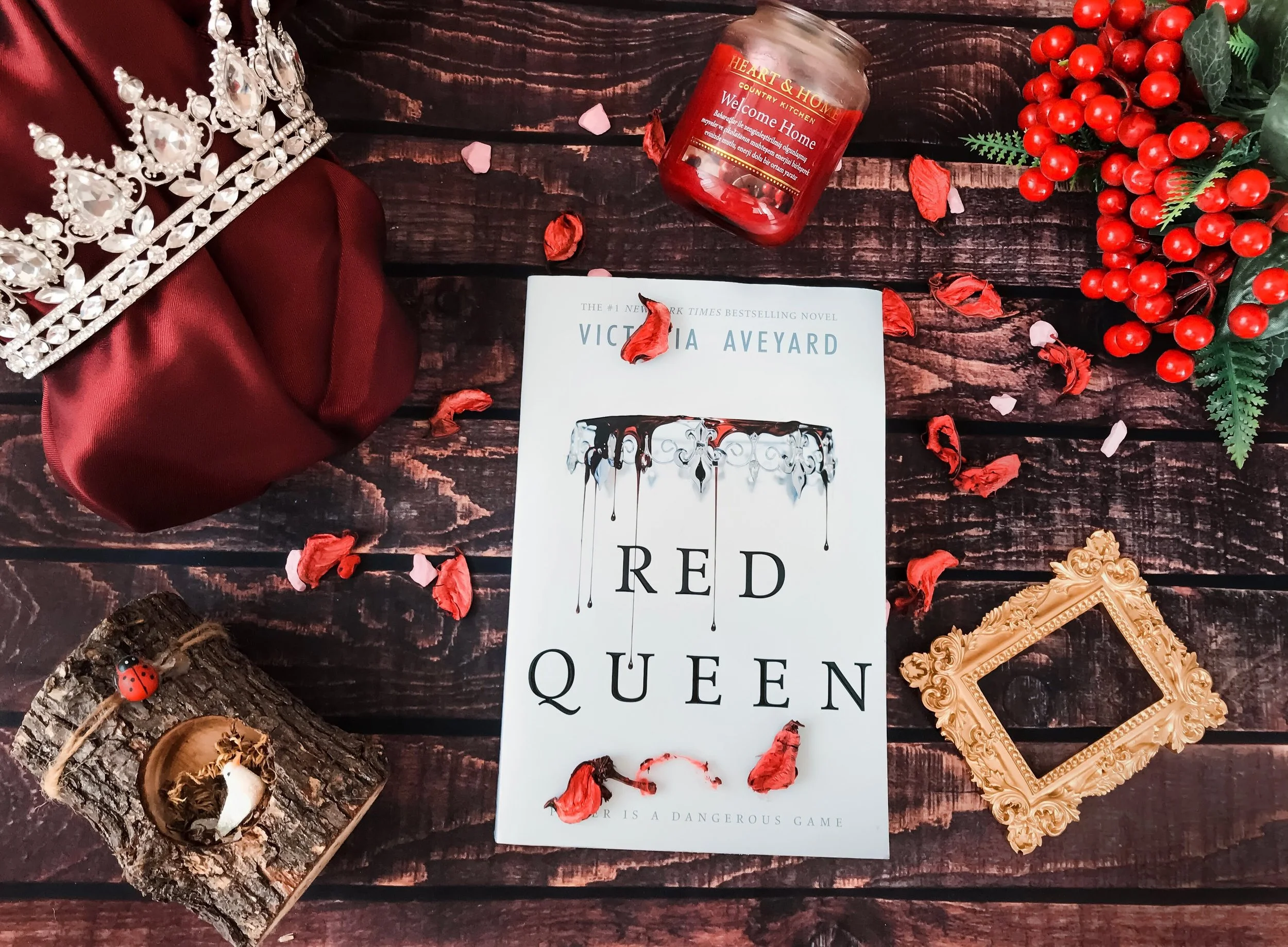 A book titled 'Red Queen' by Victoria Aveyard on a wooden surface surrounded by a red crown, a candle labeled 'Welcome Home,' a decorative golden frame, a small log with a ladybug figure, and red berries with green leaves, with scattered red and pink
