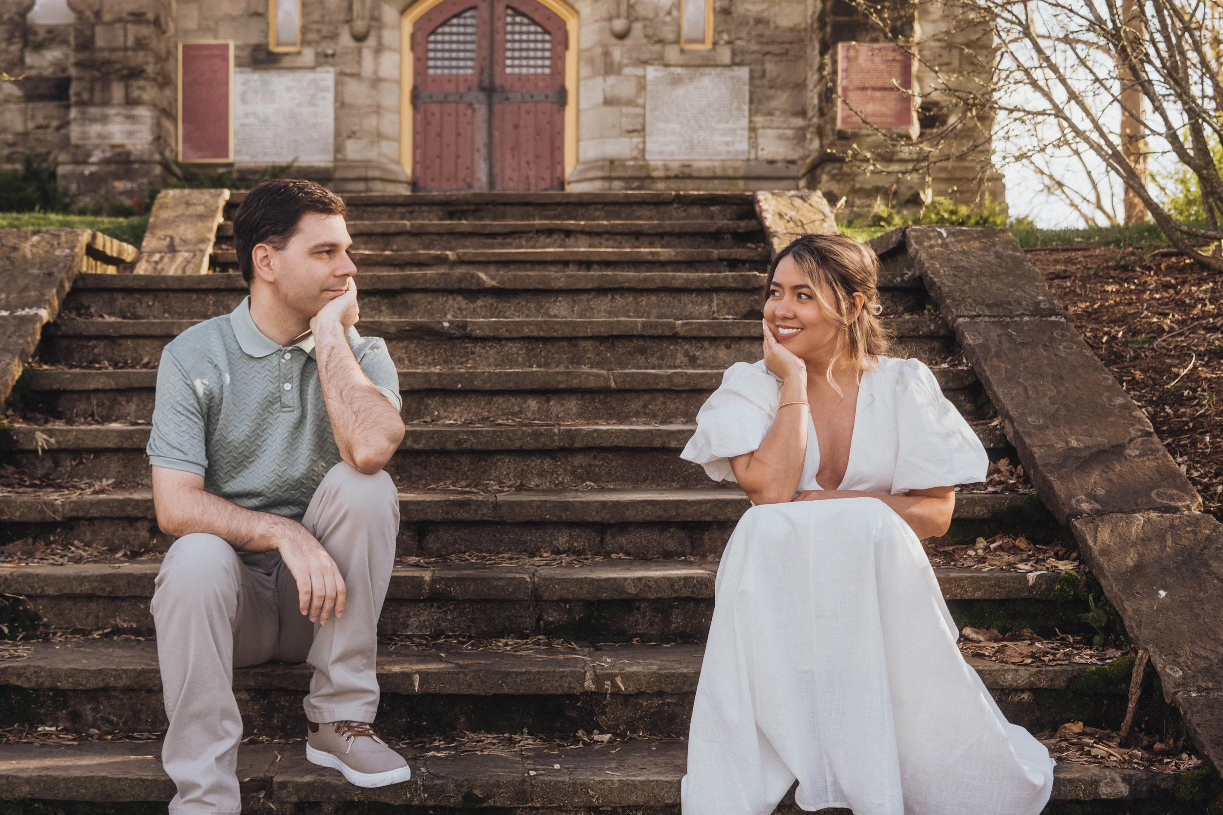 A young man and woman sitting on stone steps outside a historic stone building with a wooden arched door, having a conversation.