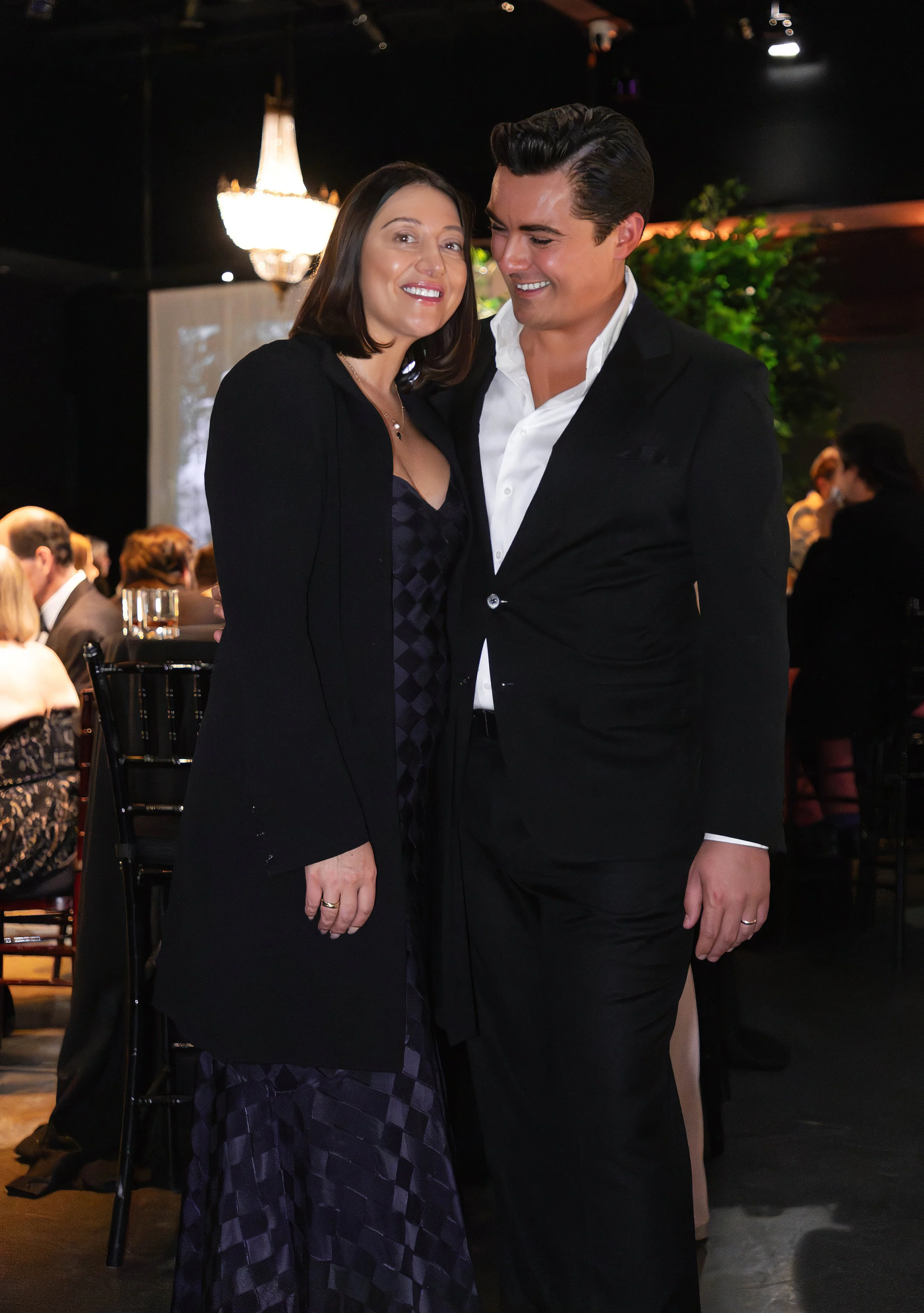 A happy couple in formal attire standing close together at an indoor event, smiling at the camera.