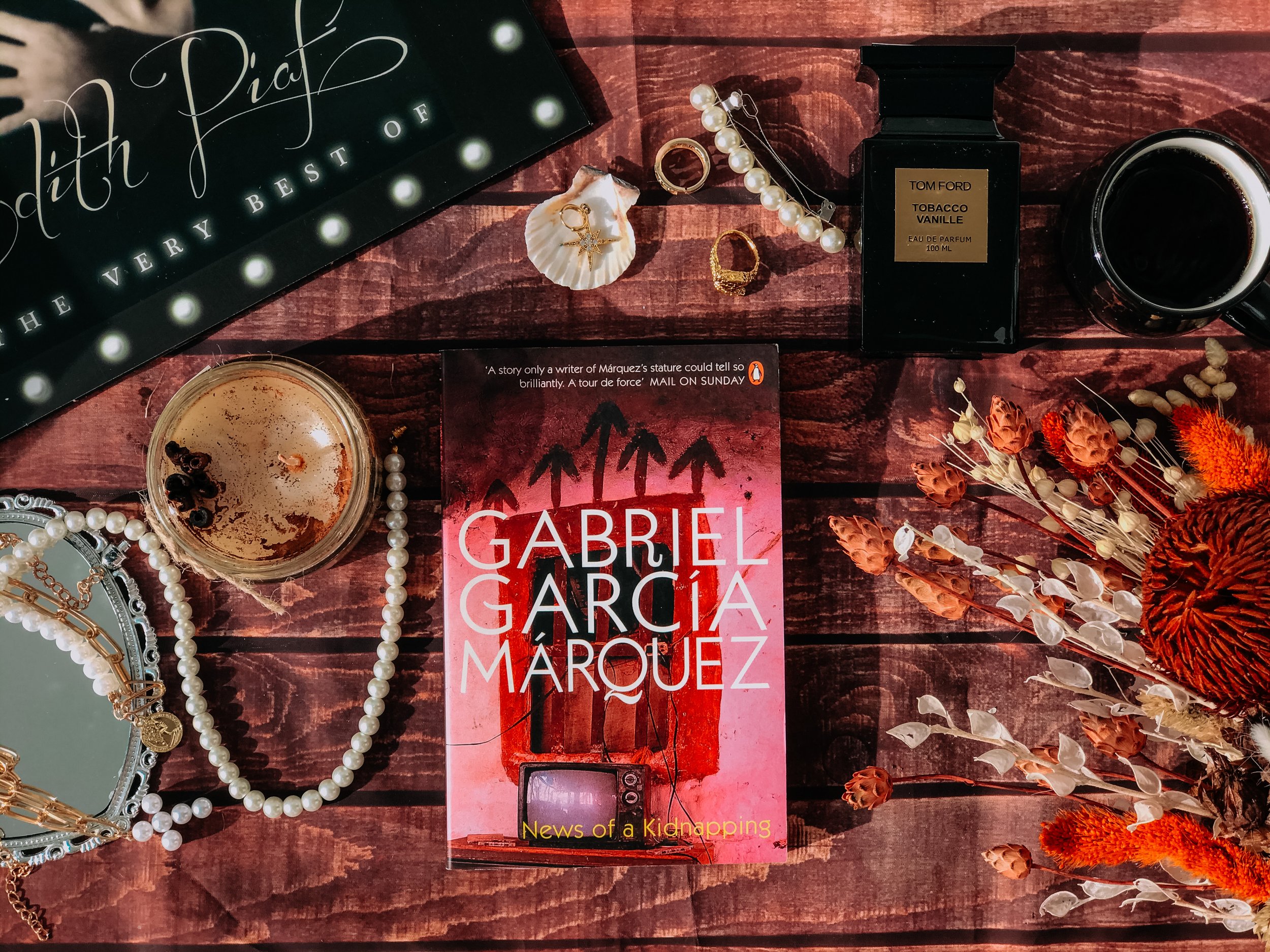 Arrangement of jewelry, a candle, a perfume bottle, a mug of coffee, a book titled "Gabriel Garcia Marquez - News of a Kidnapping", and dried flowers on a wooden surface.