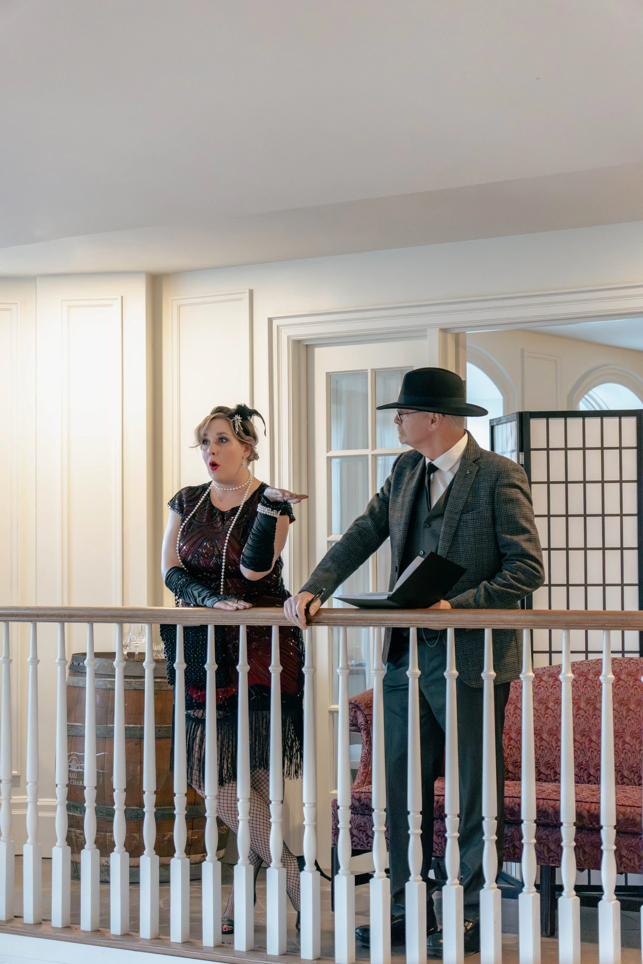 Two actors in vintage costumes perform on a balcony set for a play or film, with a woman gesturing expressive, and a man wearing a hat and holding a script or notebook.