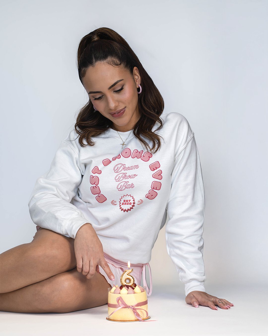 A woman with brown hair seated on the floor is celebrating her sixth birthday with a small round birthday cake decorated with pink ribbons and a candle shaped like the number 6. She is wearing a white sweatshirt with pink text and graphics and is poi