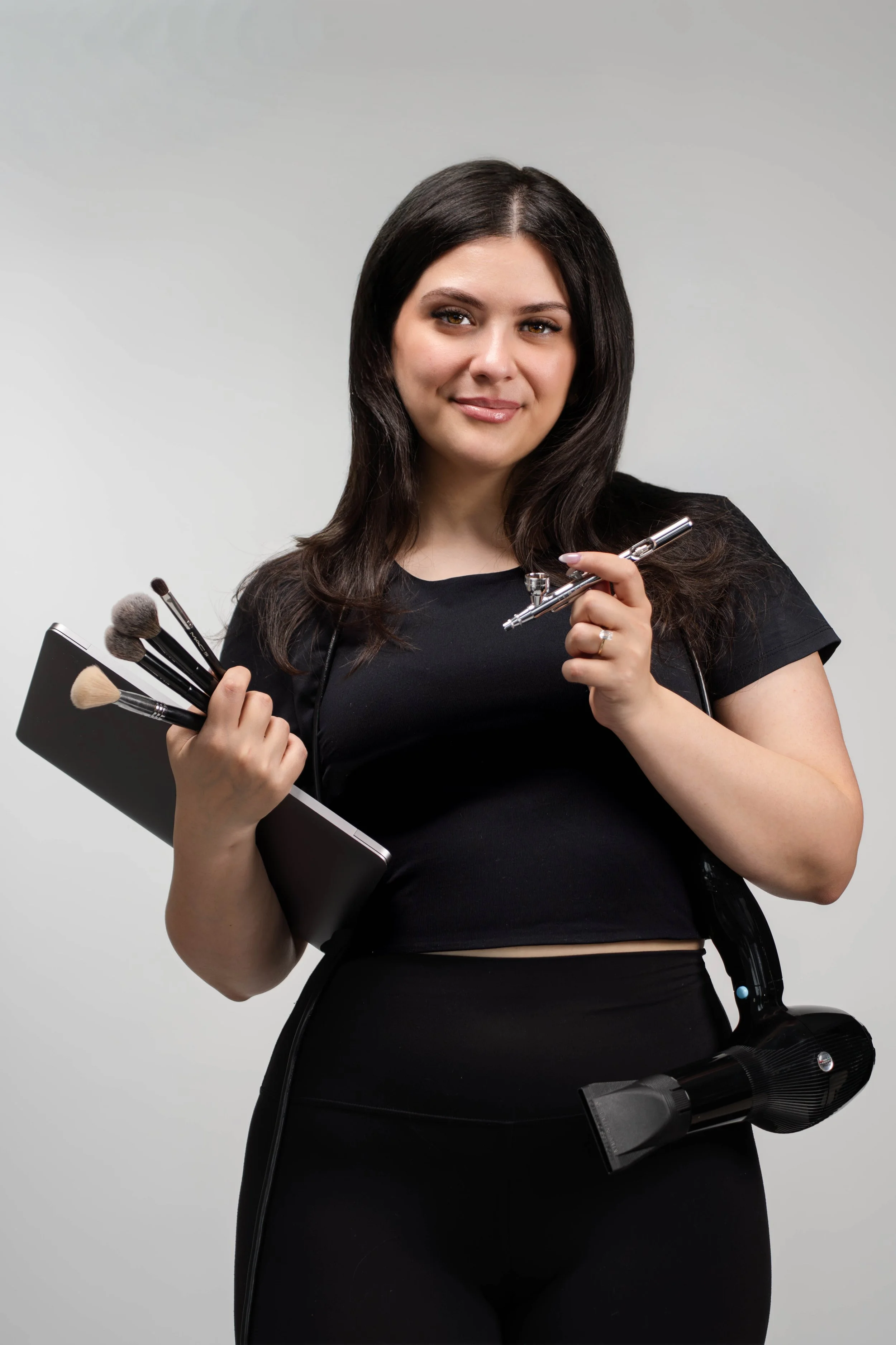 A woman holding makeup brushes, a spray gun, and a notebook, dressed in black, standing against a plain background.