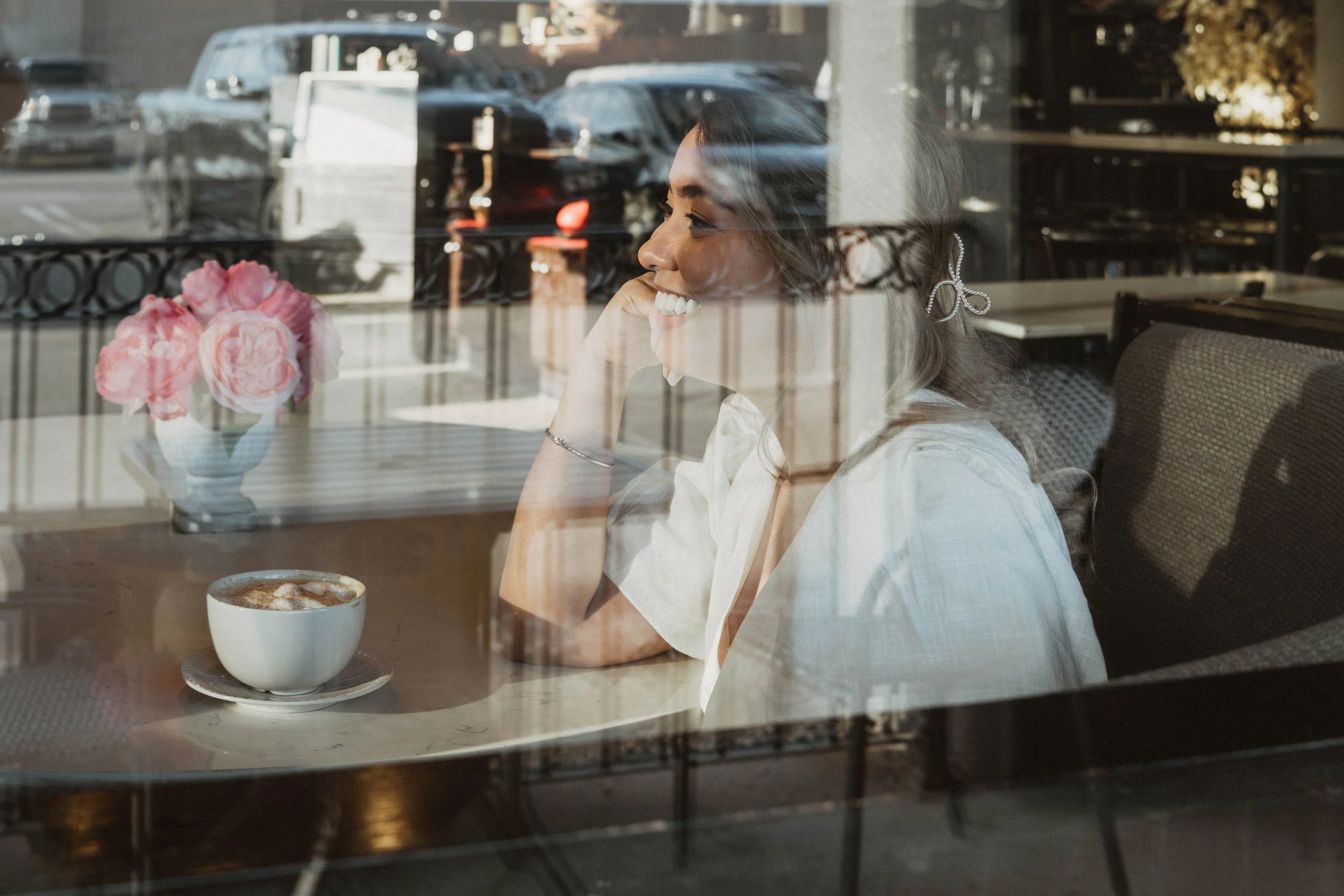A woman sitting inside a cafe, smiling and resting her chin on her hand, seen through a window with reflections of parked cars outside. On the table, there is a cup of coffee and a vase with pink flowers.