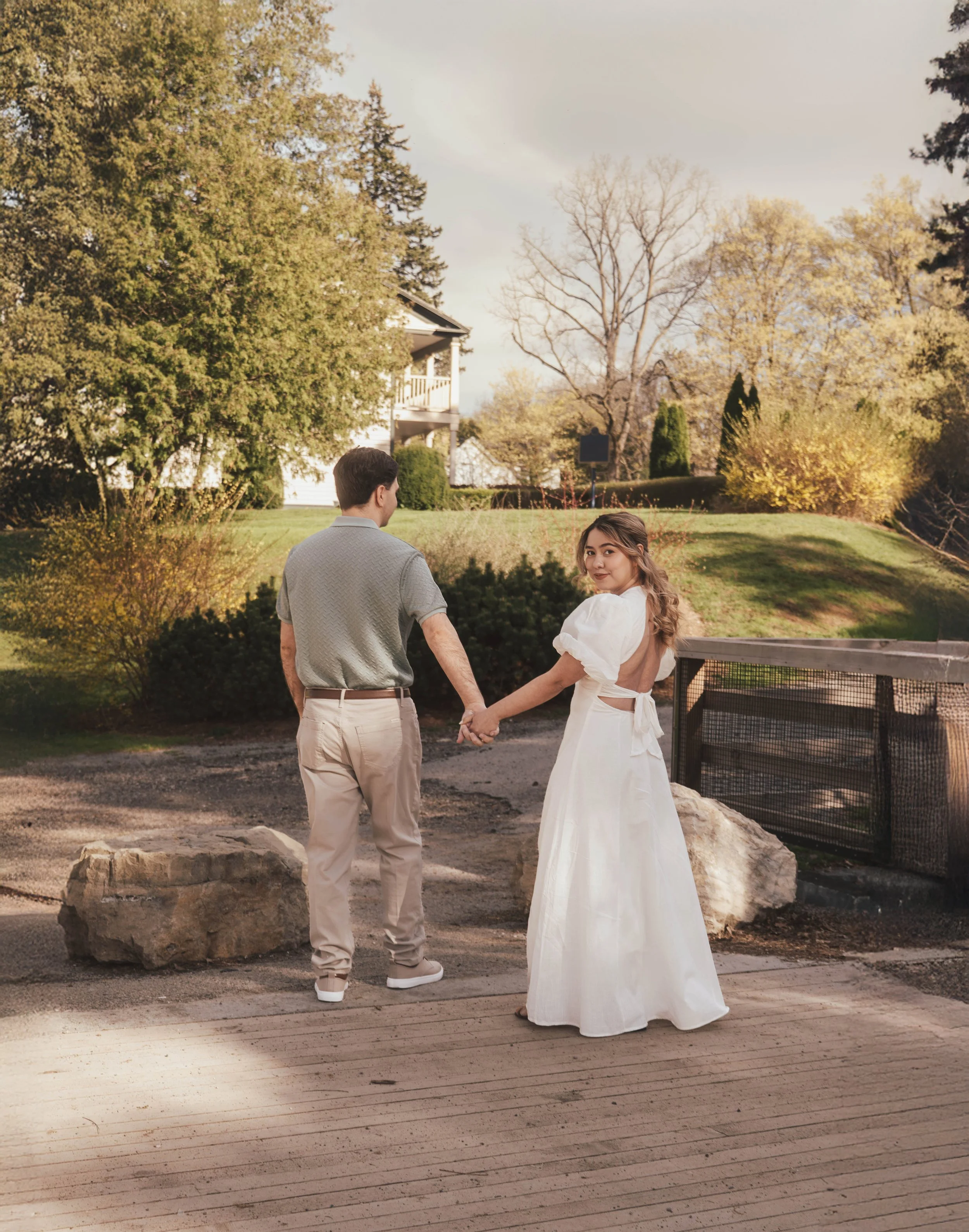 A couple holding hands outdoors in a park-like setting during autumn. The woman wears a white dress, and the man wears a light shirt and beige pants. Trees with yellowing leaves are in the background, and a house is visible behind the trees.
