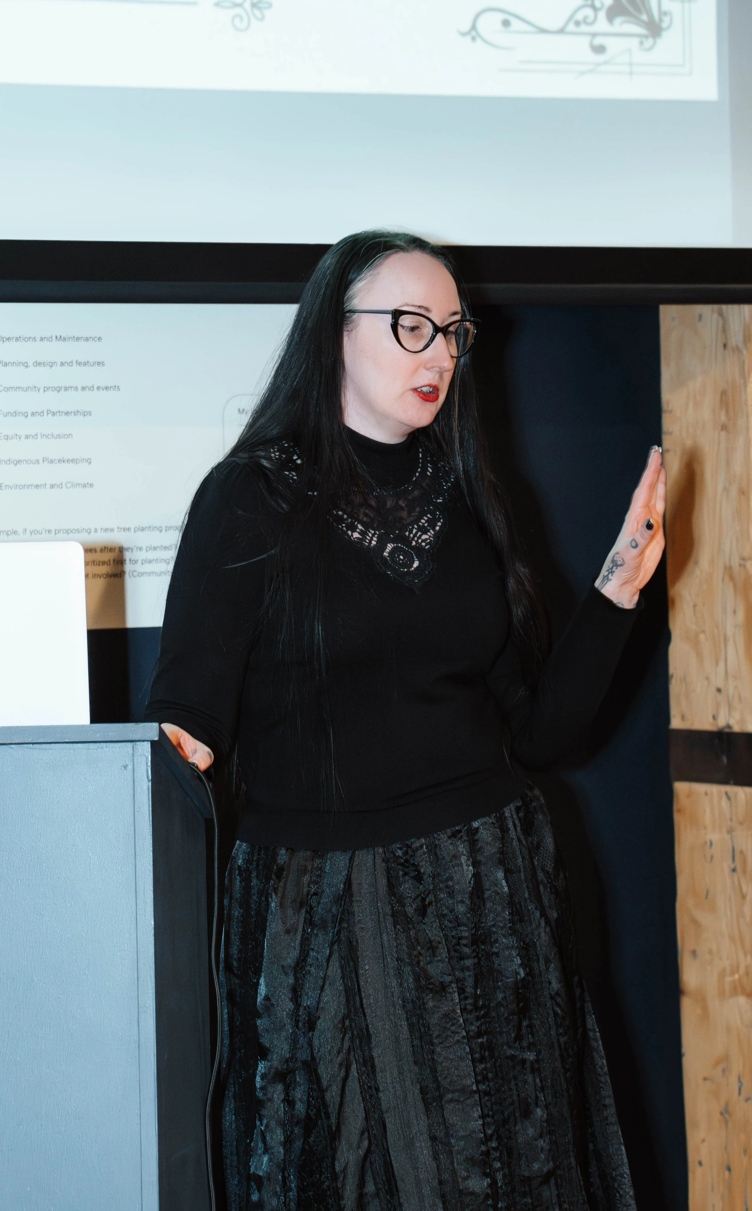 A woman with long black hair and glasses standing beside a podium, gesturing with her right hand, during a presentation with a slide displayed on the wall behind her.