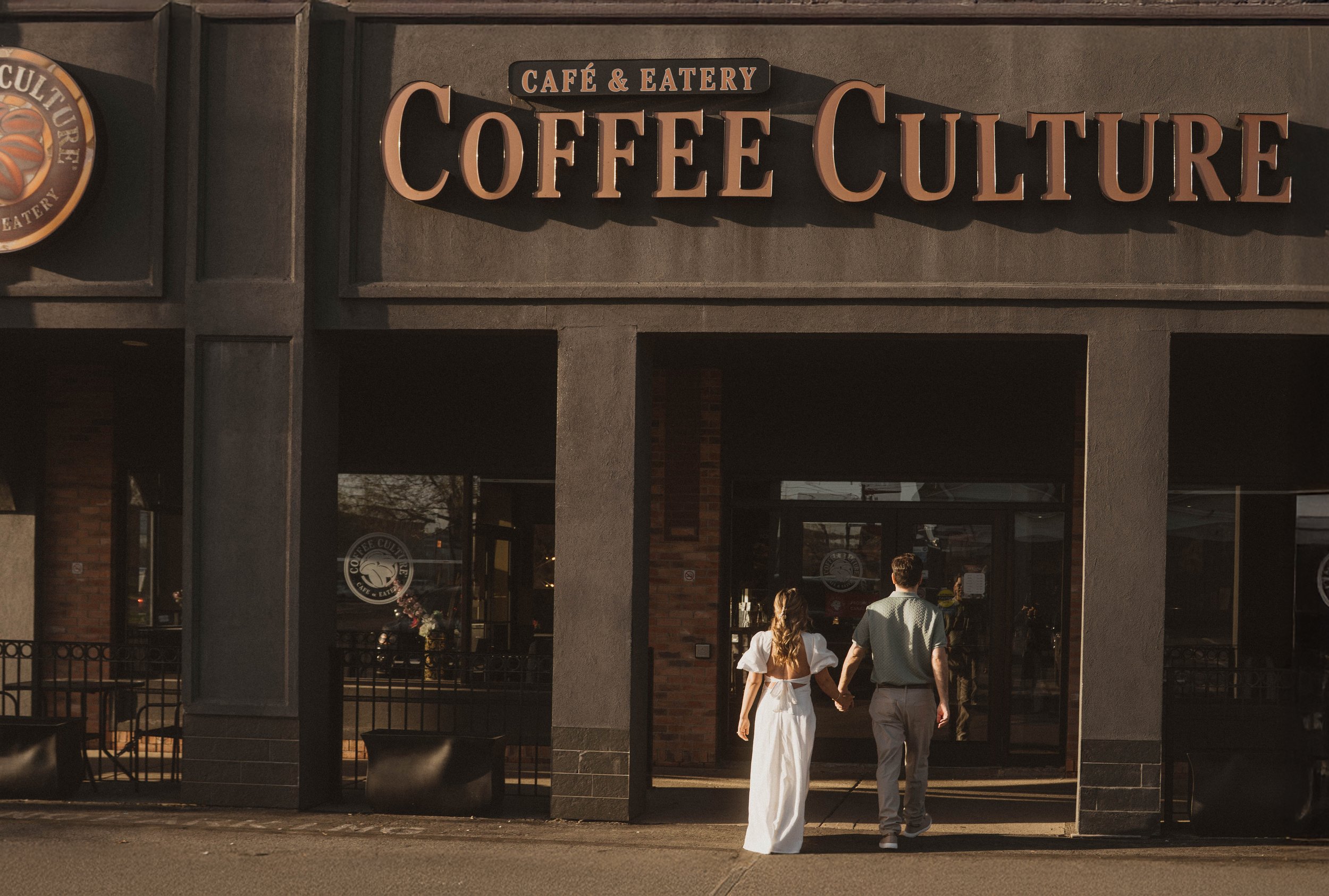 The storefront of a café and eatery named 'Coffee Culture' with a large sign above the entrance. Two people, a woman in a white dress and a man in a short-sleeved shirt and pants, are walking hand in hand towards the building.