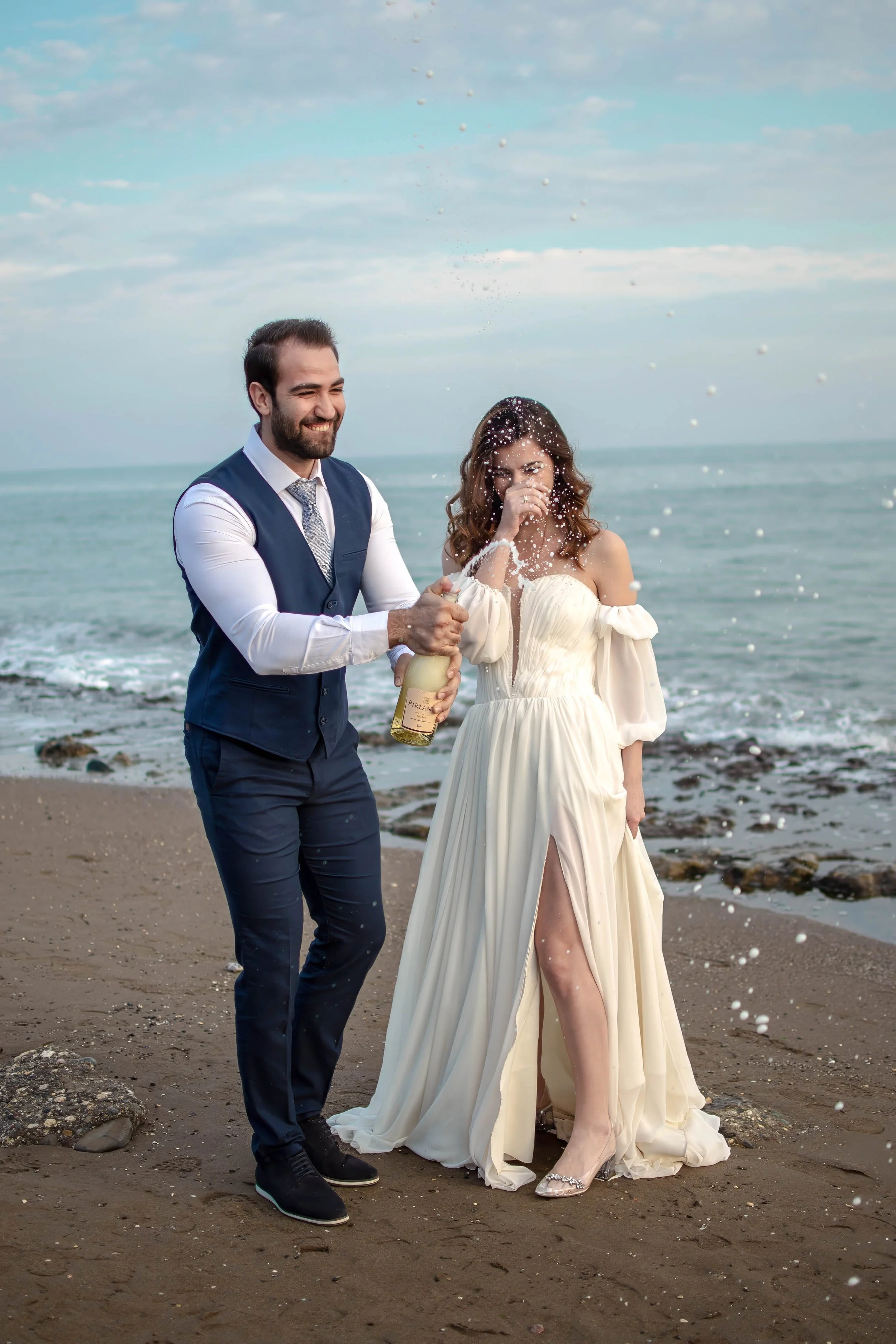 A couple in wedding attire celebrating on a beach, with the man spraying champagne on the woman as they smile and enjoy the moment.