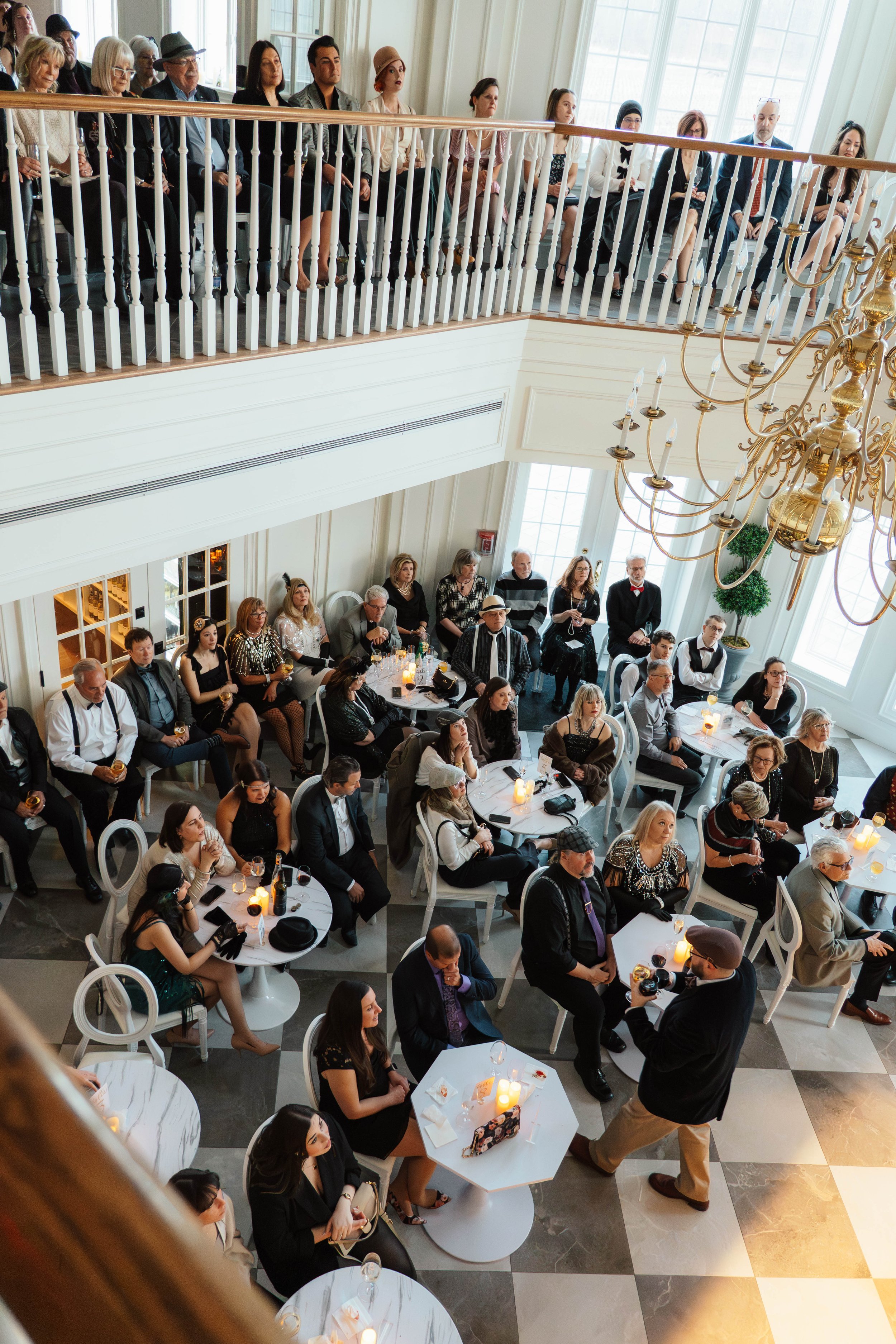 Guests seated at tables during an indoor event or reception, with some standing on a balcony overlooking the scene, in a well-lit room with large windows and a chandelier.