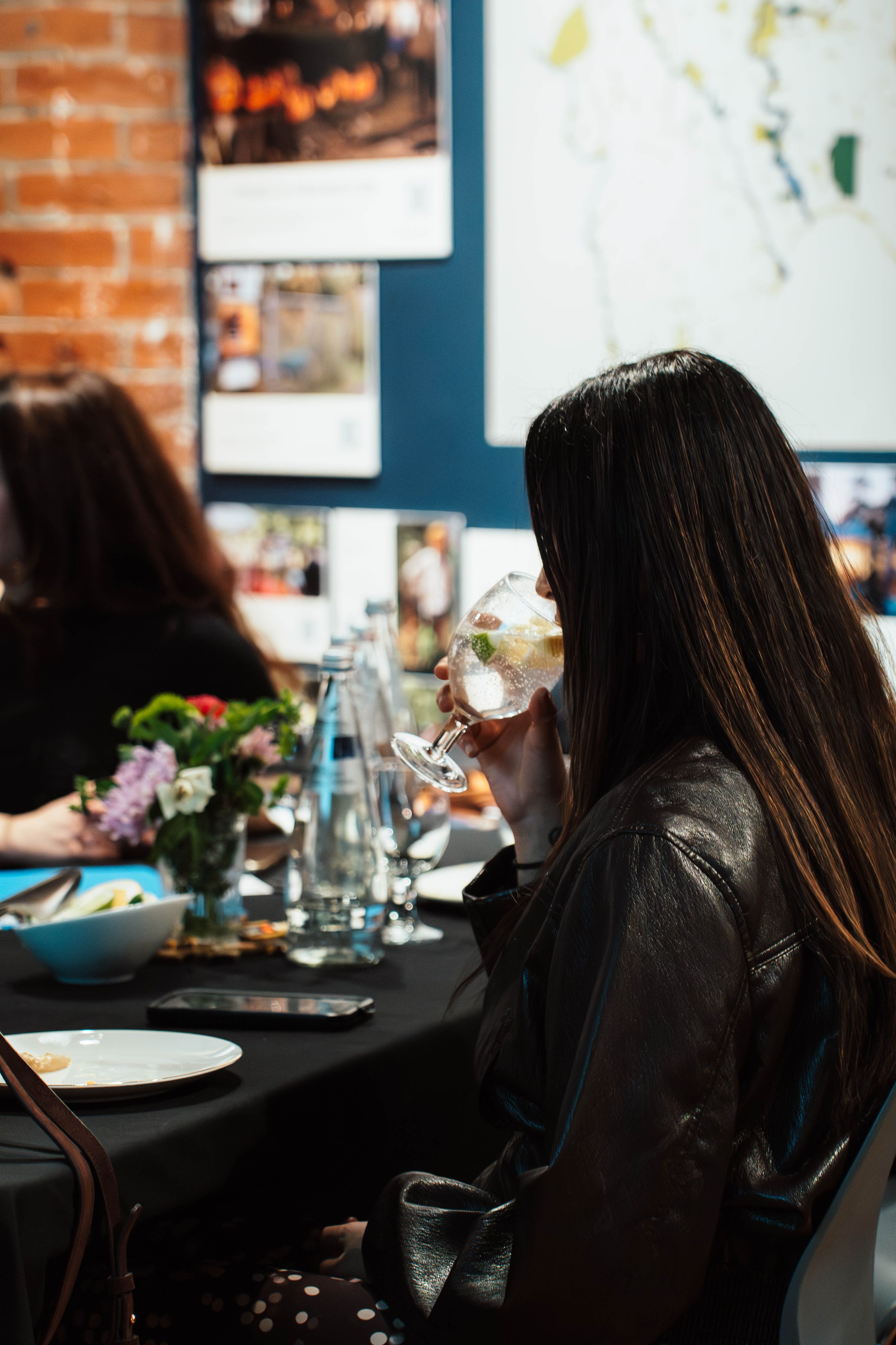 A woman with long dark hair sitting at a table in a restaurant, drinking a cocktail with lemon and lime slices, with a flower centerpiece, and other people and photos on the wall in the background.
