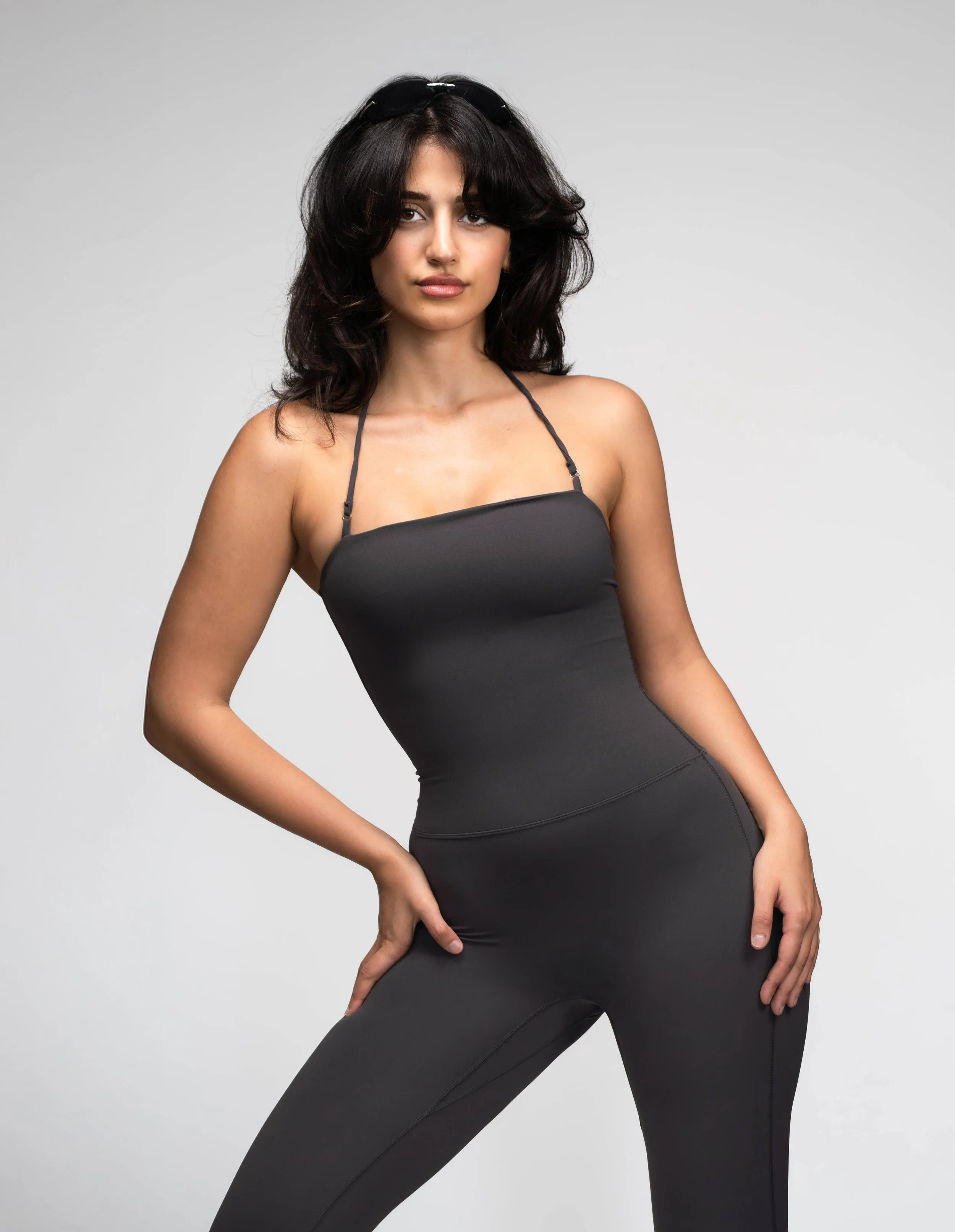 Young woman with dark hair wearing a black halter top and matching black pants posing against a gray background.