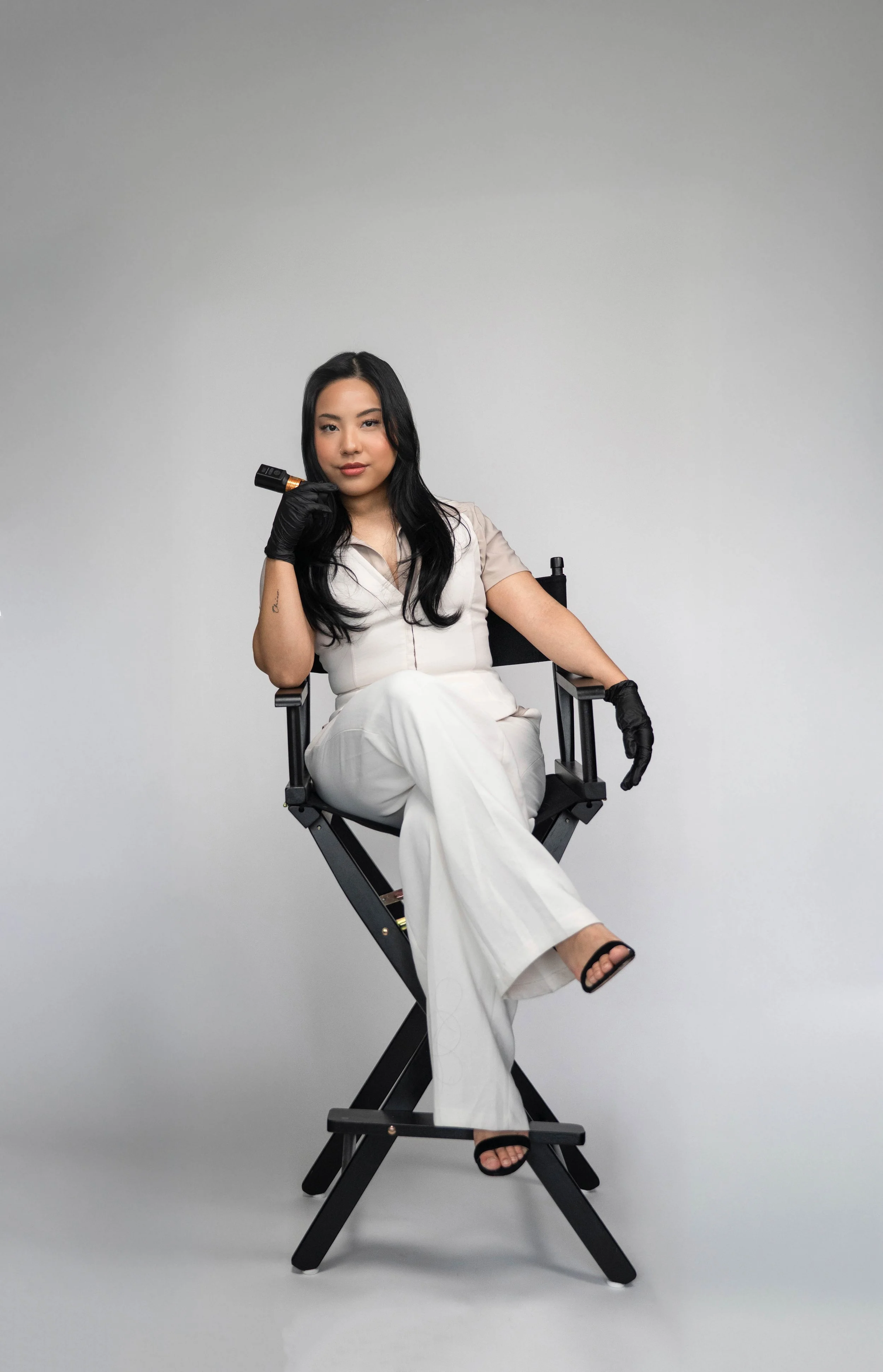 A woman with long black hair, wearing black gloves, beige top, white pants, and black sandals, sits in a director's chair holding a tattoo machine, against a plain gray background.