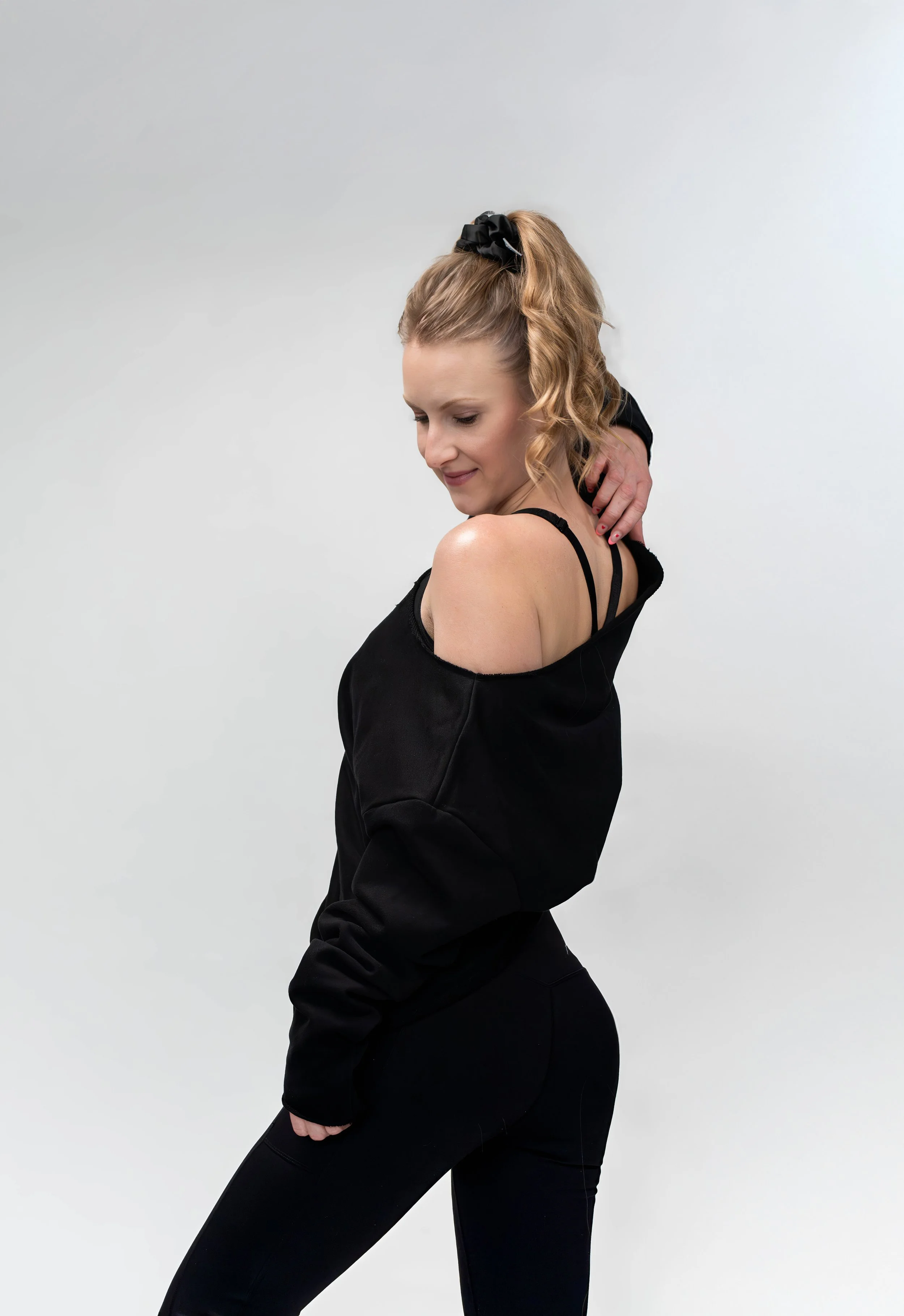 A woman with blonde curly hair tied back with a black scrunchie, wearing a black off-the-shoulder top and black leggings, standing against a plain light gray background.