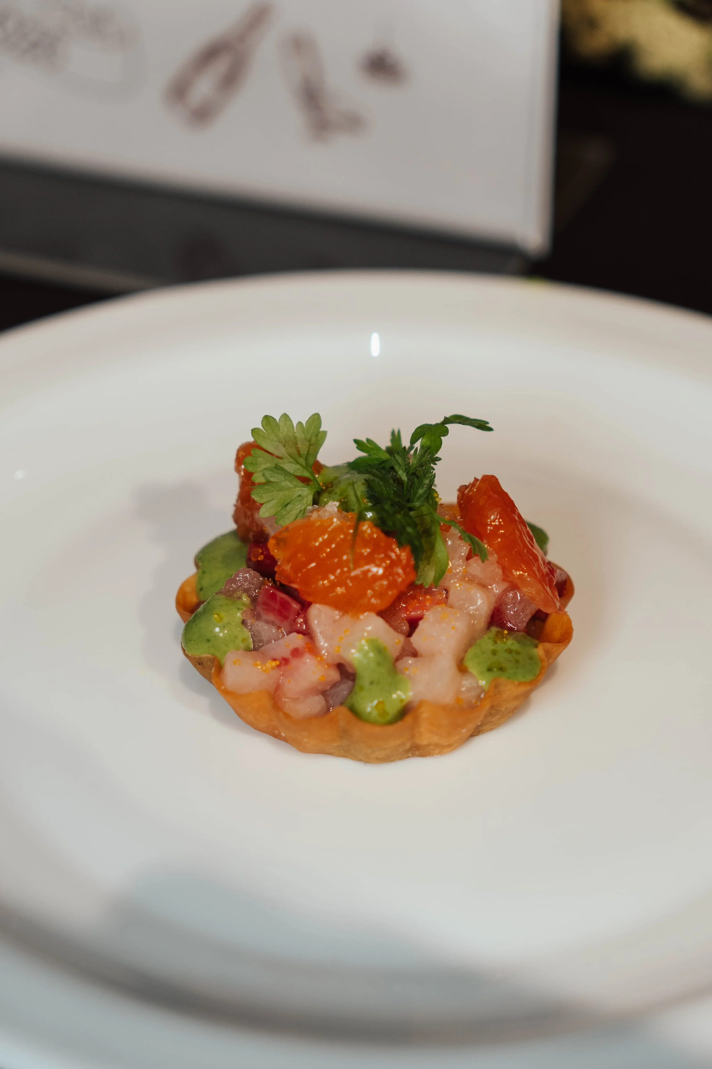 A small, round tart topped with chopped raw seafood, drizzled green sauce, and garnished with a sprig of cilantro.