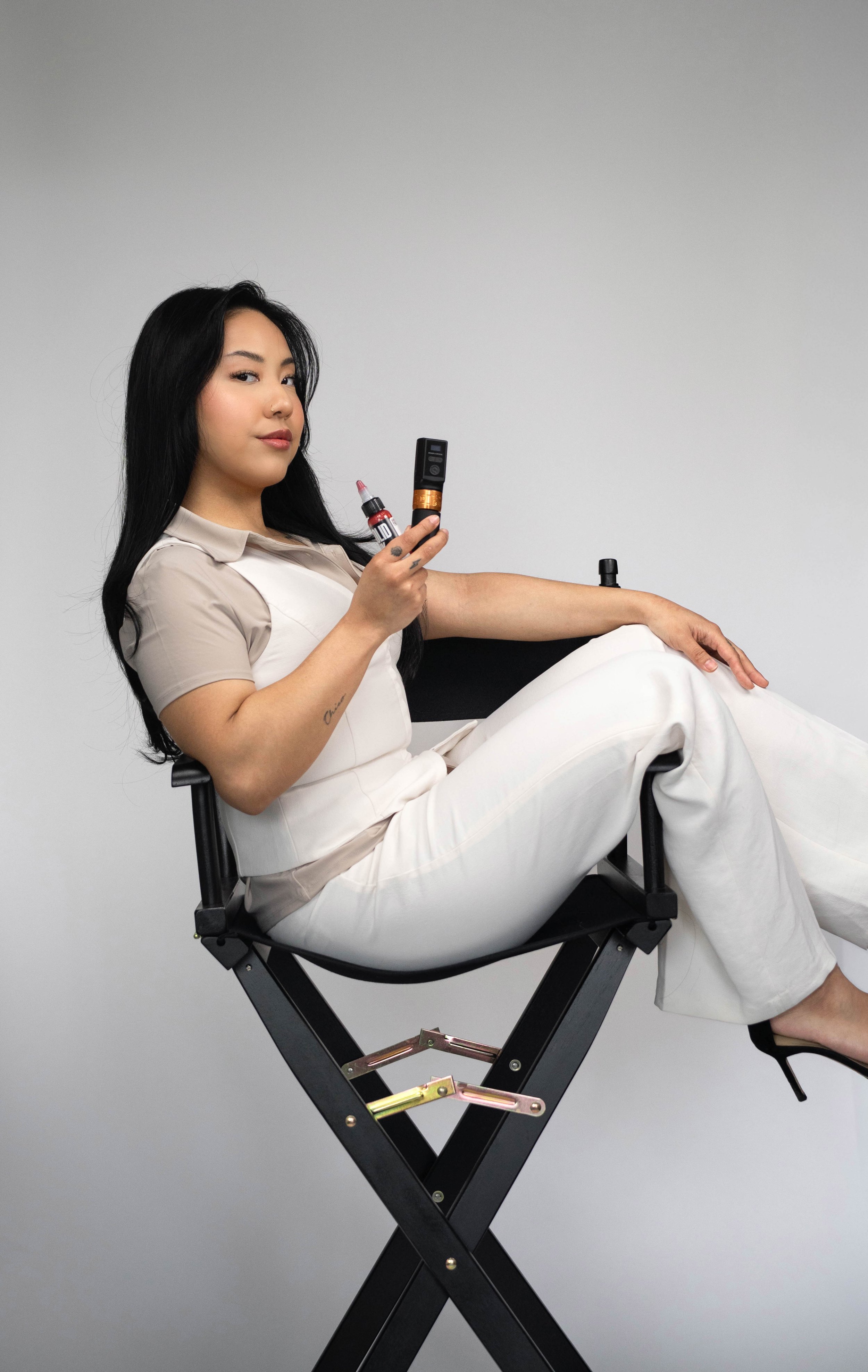 Young woman sitting on a director's chair, holding a vape and a vape juice bottle, wearing a beige and white outfit, with a neutral background.