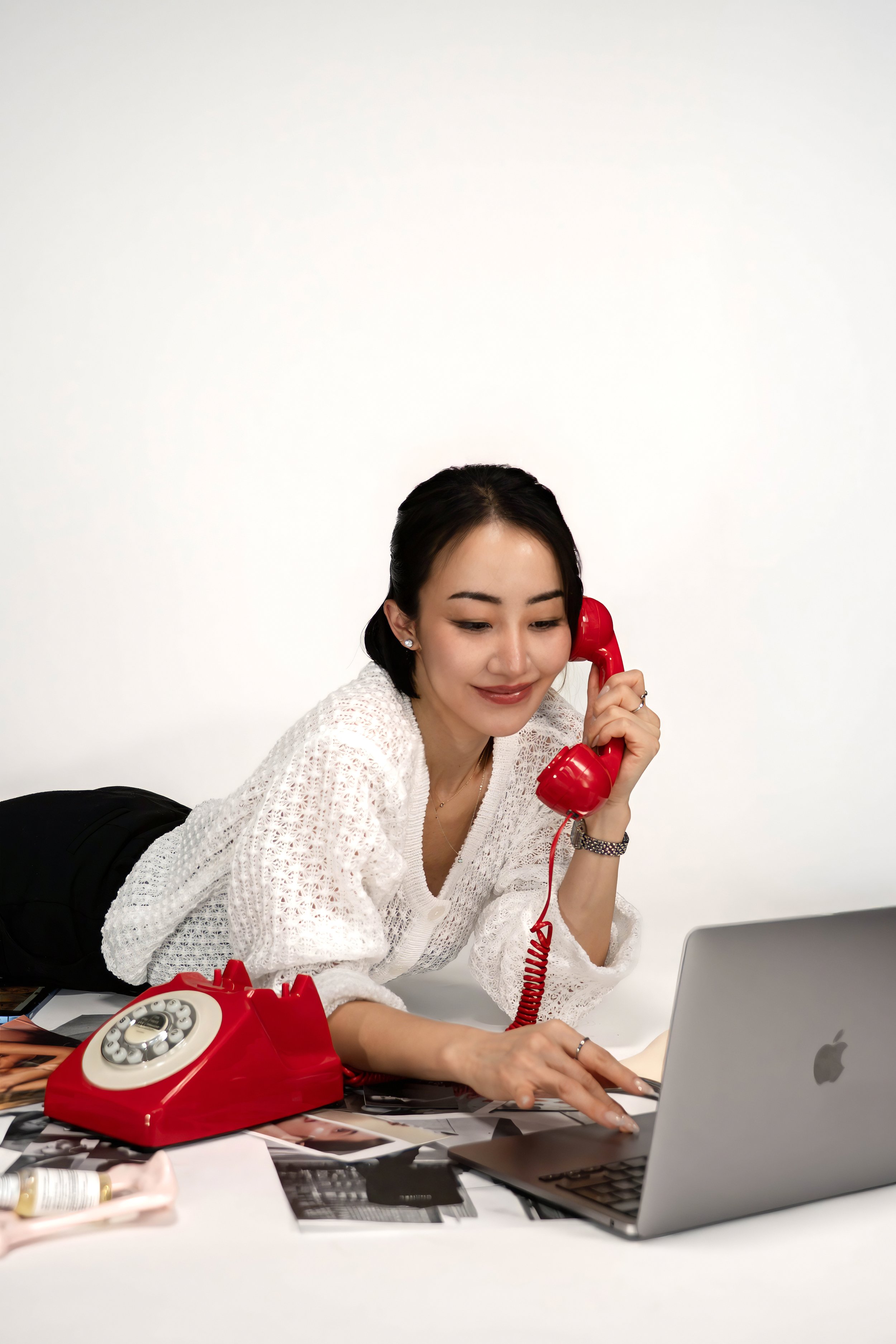 Woman lying on her stomach talking on a red rotary phone, working on a laptop with scattered magazines and photos on a white table.