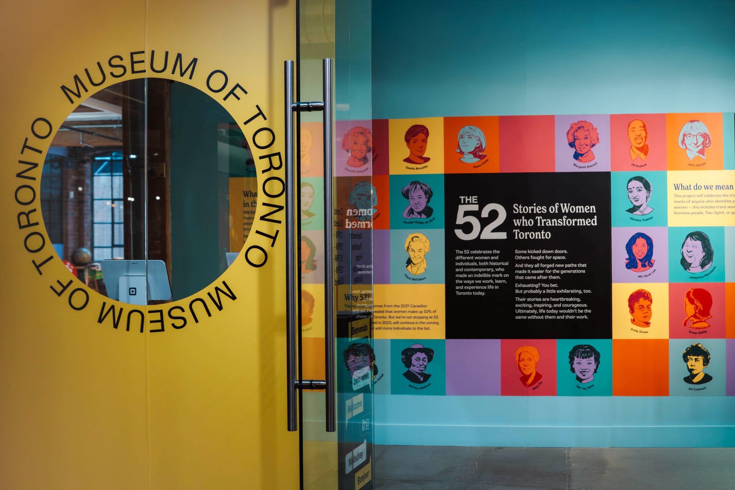 Entrance to the Toronto Museum of Tolerance featuring a glass door with yellow frame and black text, and a colorful wall display celebrating 52 stories of women who transformed Toronto, with illustrated portraits of women and informational text.
