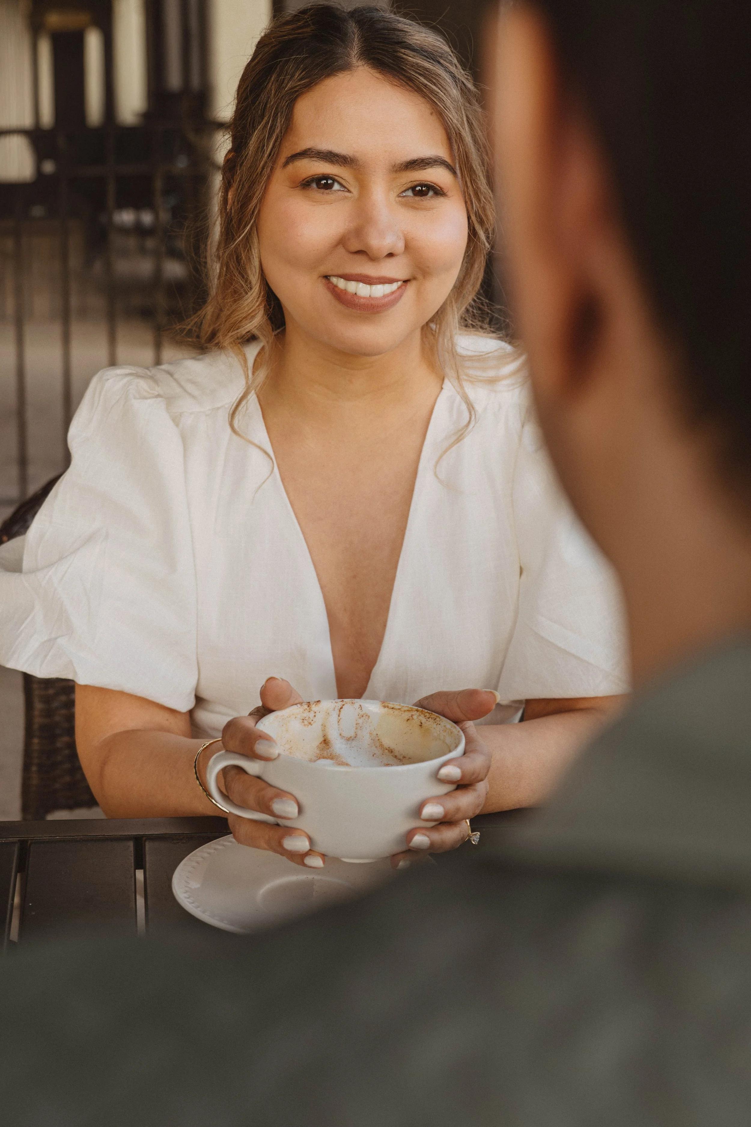 A smiling woman with wavy brown hair wearing a white blouse, holding a white mug with coffee and cinnamon, sitting at a table in a cozy cafe, engaging in conversation with someone.