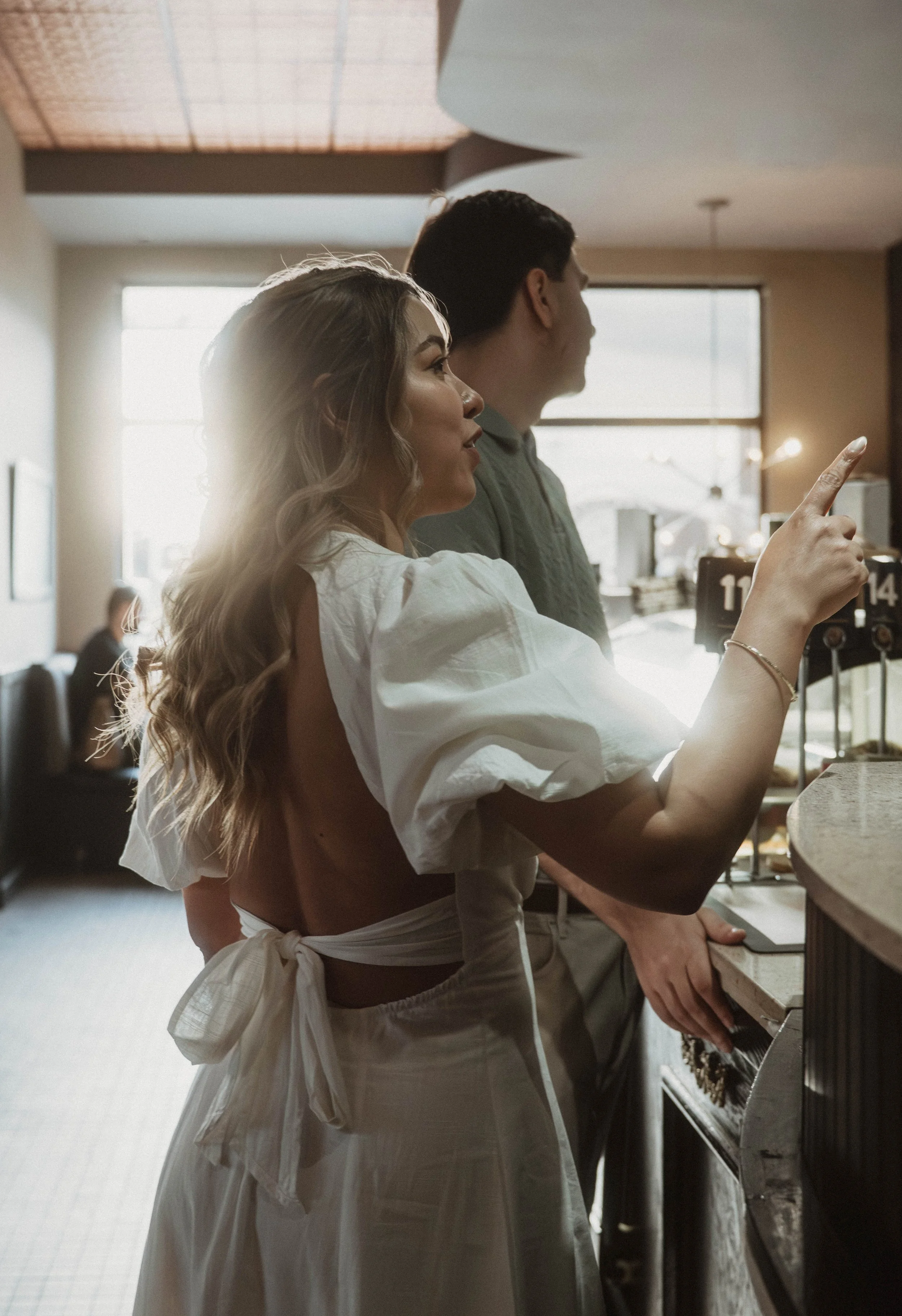 A woman with long, wavy hair wearing a white dress and apron, and a man with short dark hair in a green shirt, are standing at a counter in a cafe. The woman is pointing at an order number sign, and they are both looking in the same direction. The su