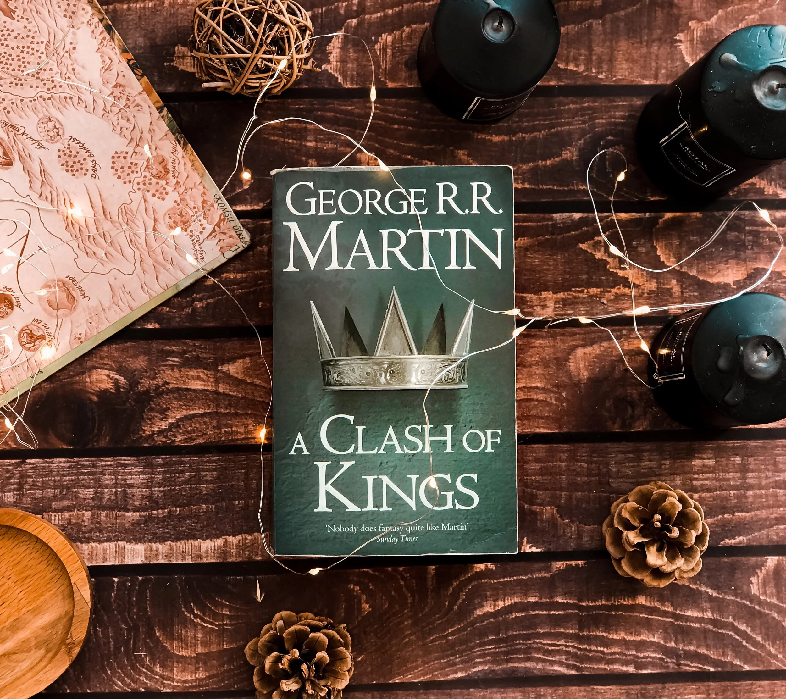 A book titled 'A Clash of Kings' by George R.R. Martin on a wooden surface, surrounded by pine cones, string lights, a portion of a map, and black candles.