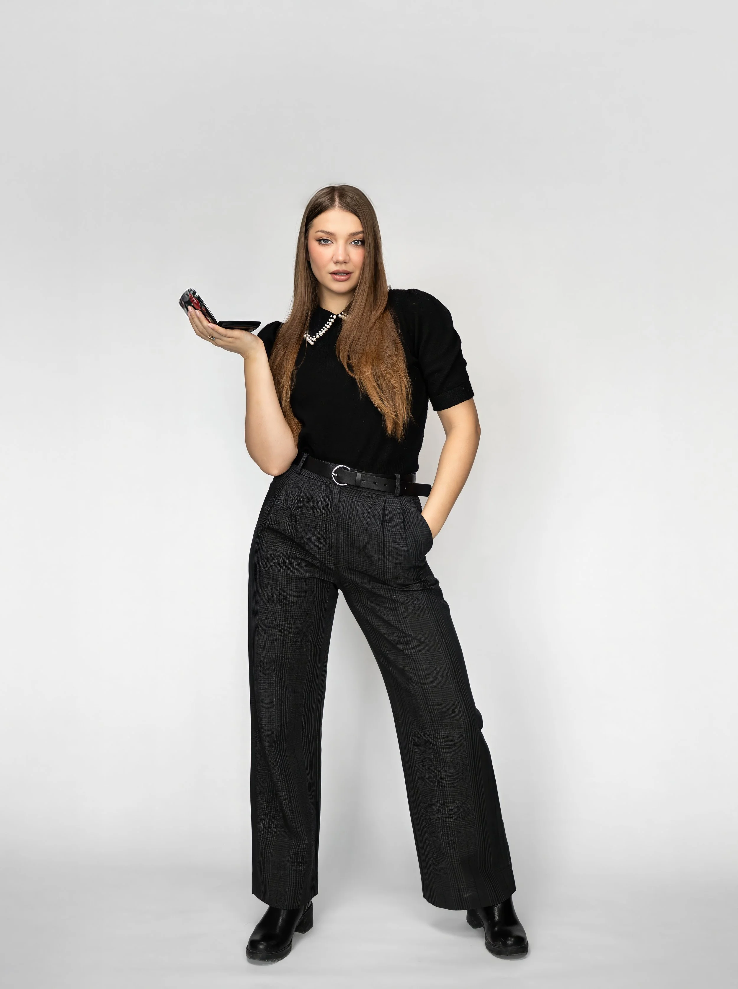 A young woman with long brown hair wearing a black shirt, plaid black trousers, black boots, and a pearl necklace, standing against a plain light gray background. She holds a smartphone in her right hand and has her left hand in her pocket.