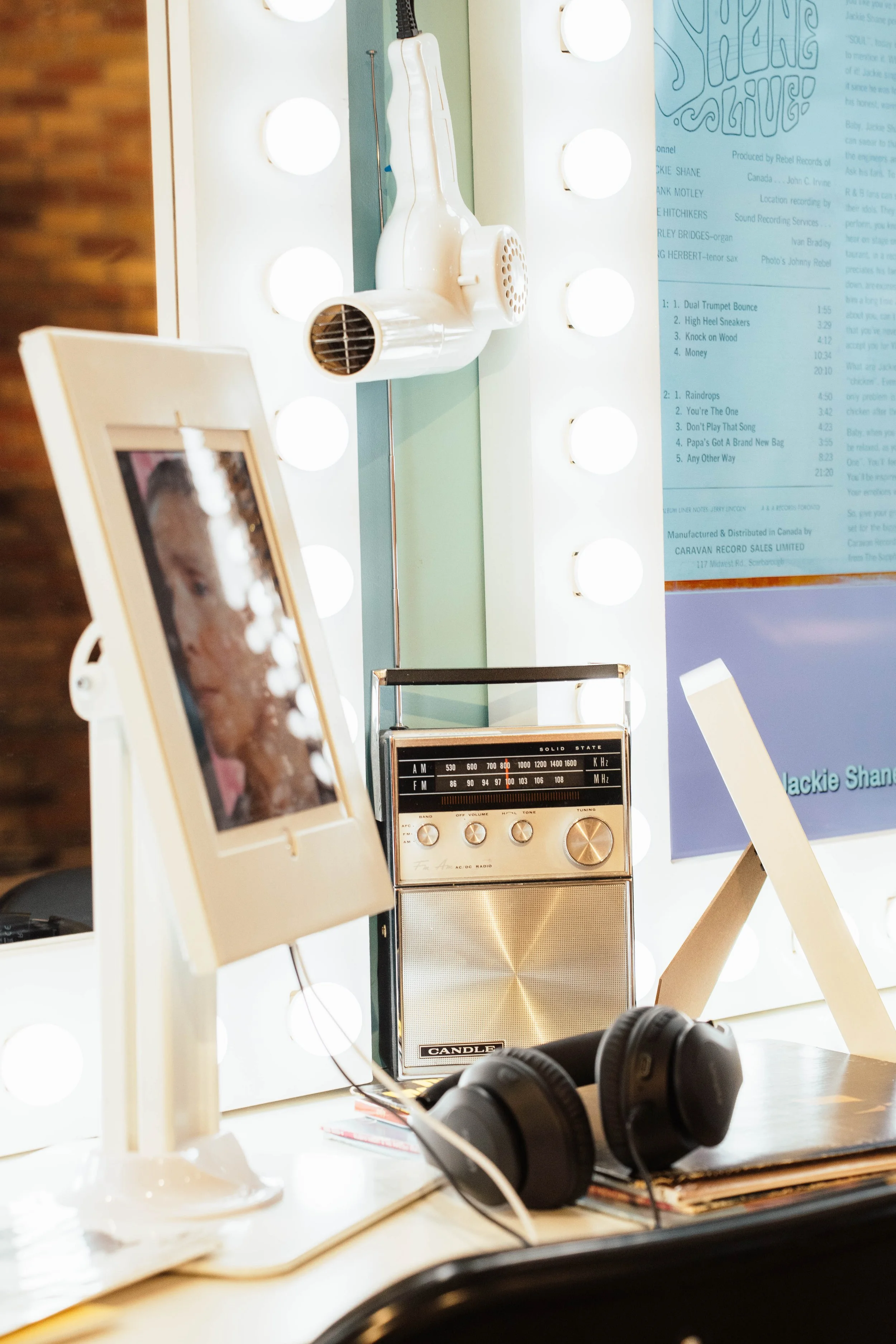 A vanity table with a mirror surrounded by round light bulbs, a vintage radio, a pair of black headphones, a framed photograph, and a blue poster or menu in the background.